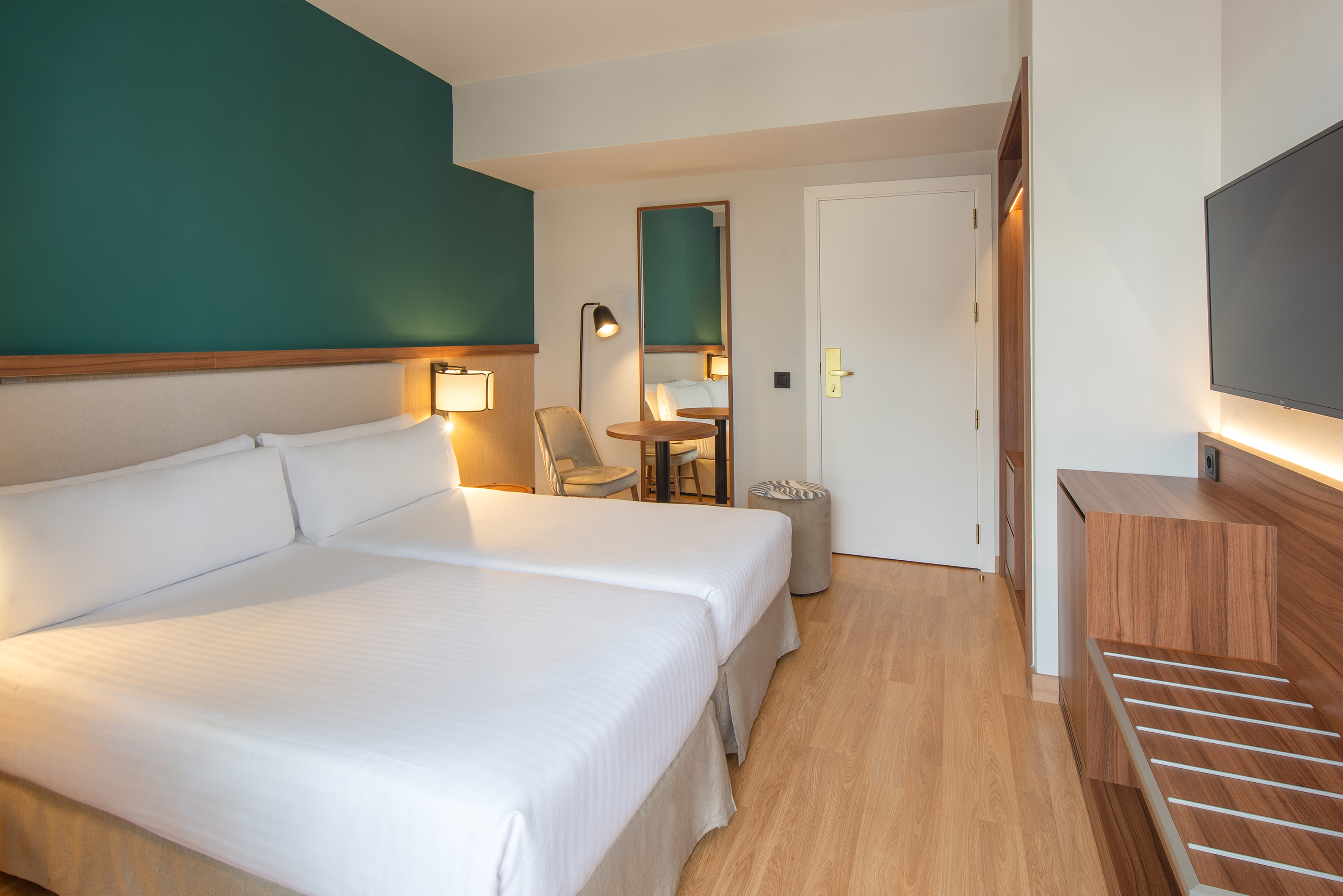 Modern hotel room with twin beds, teal wall, wood accents, TV, and luggage rack.