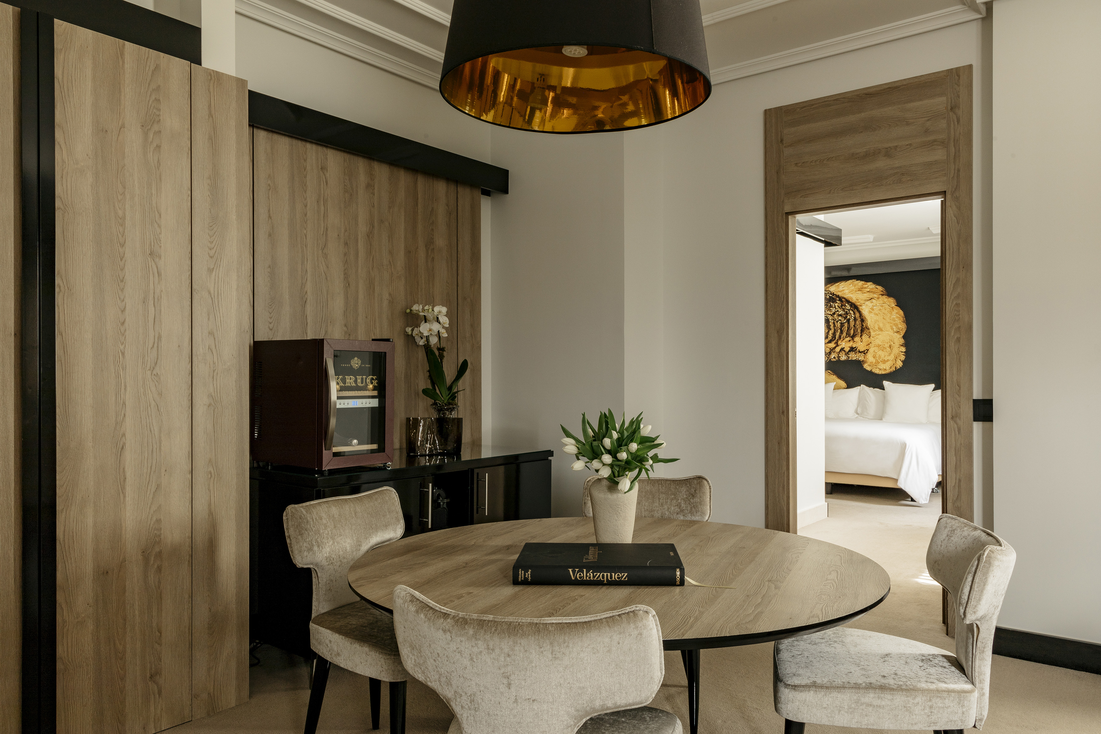 Modern hotel suite dining area with round wooden table, minibar, and bedroom view.