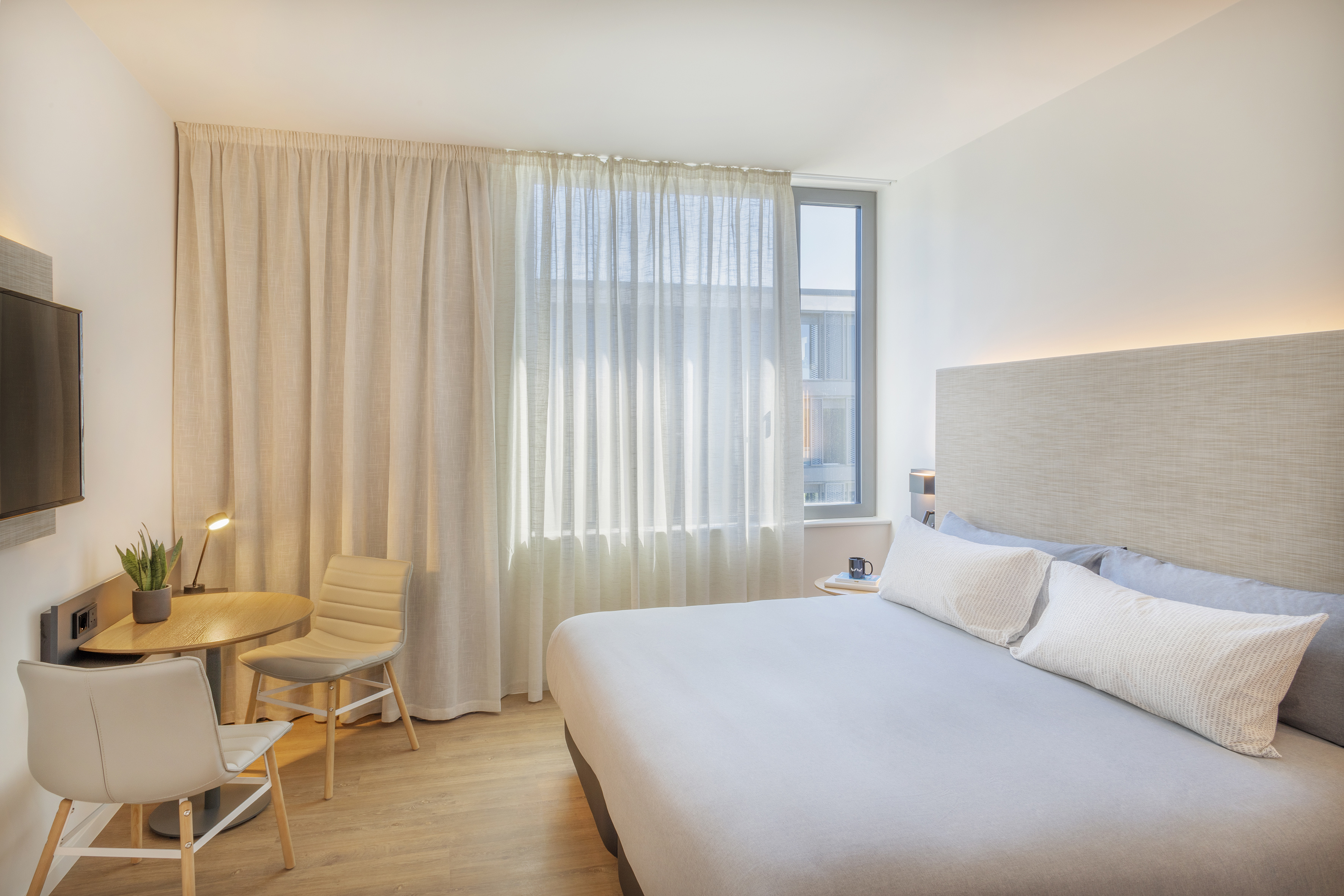 Modern hotel room with queen bed, desk, chairs, TV, and sunlit window.