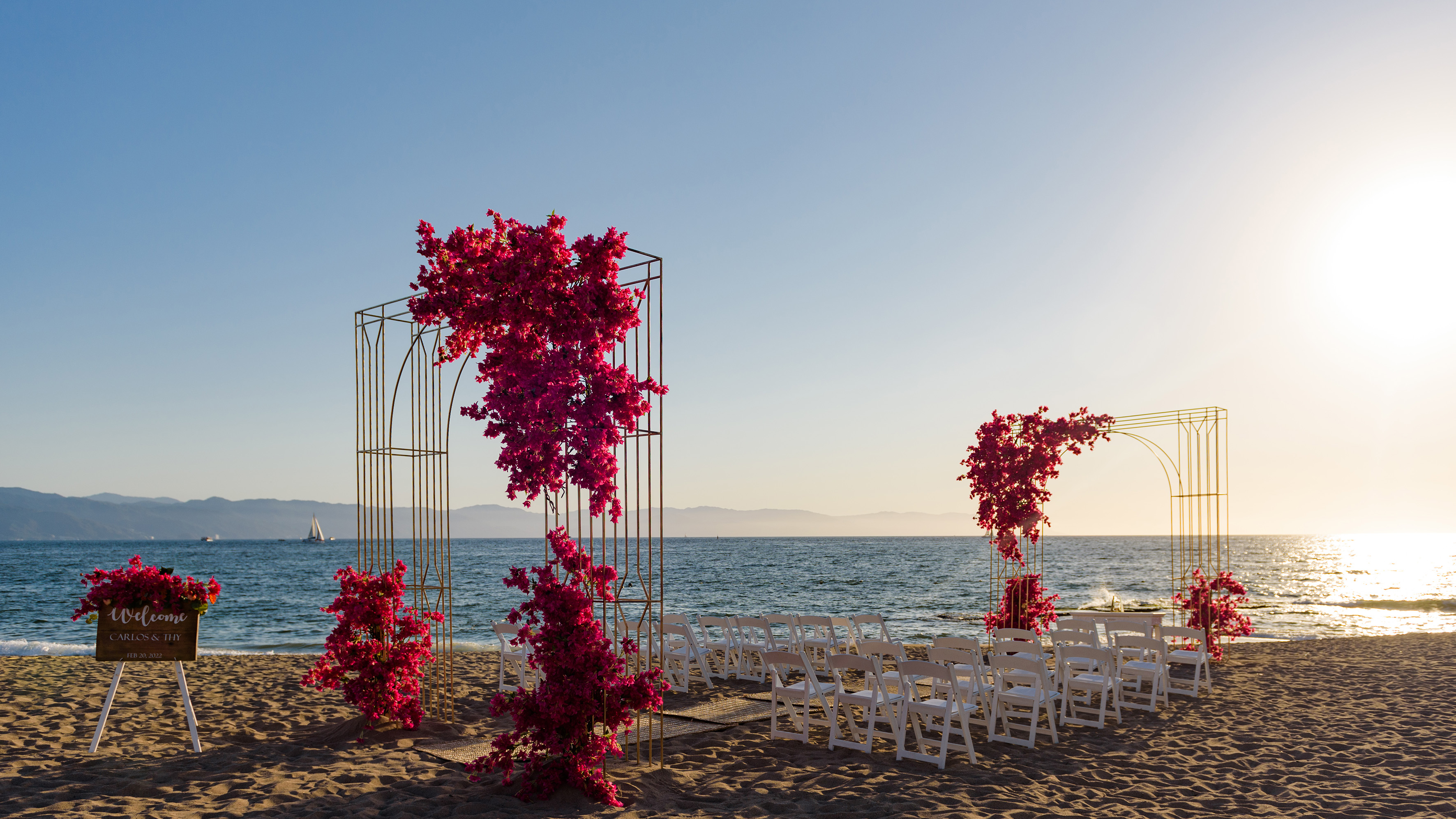 Bougainvillea wedding arches, white chairs on a sandy beach at sunset.
