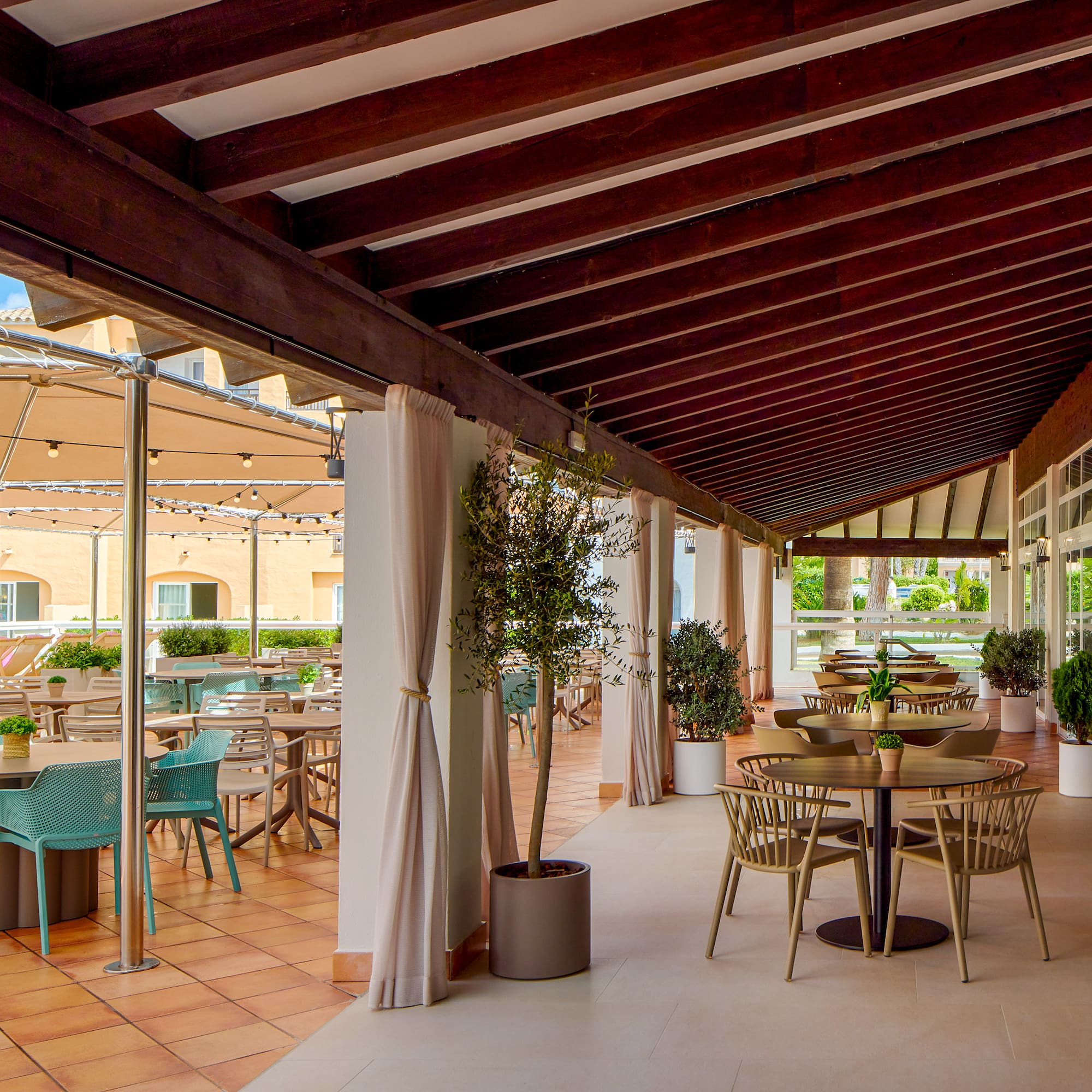 Outdoor resort dining terrace with wooden beams, various tables, and plants.