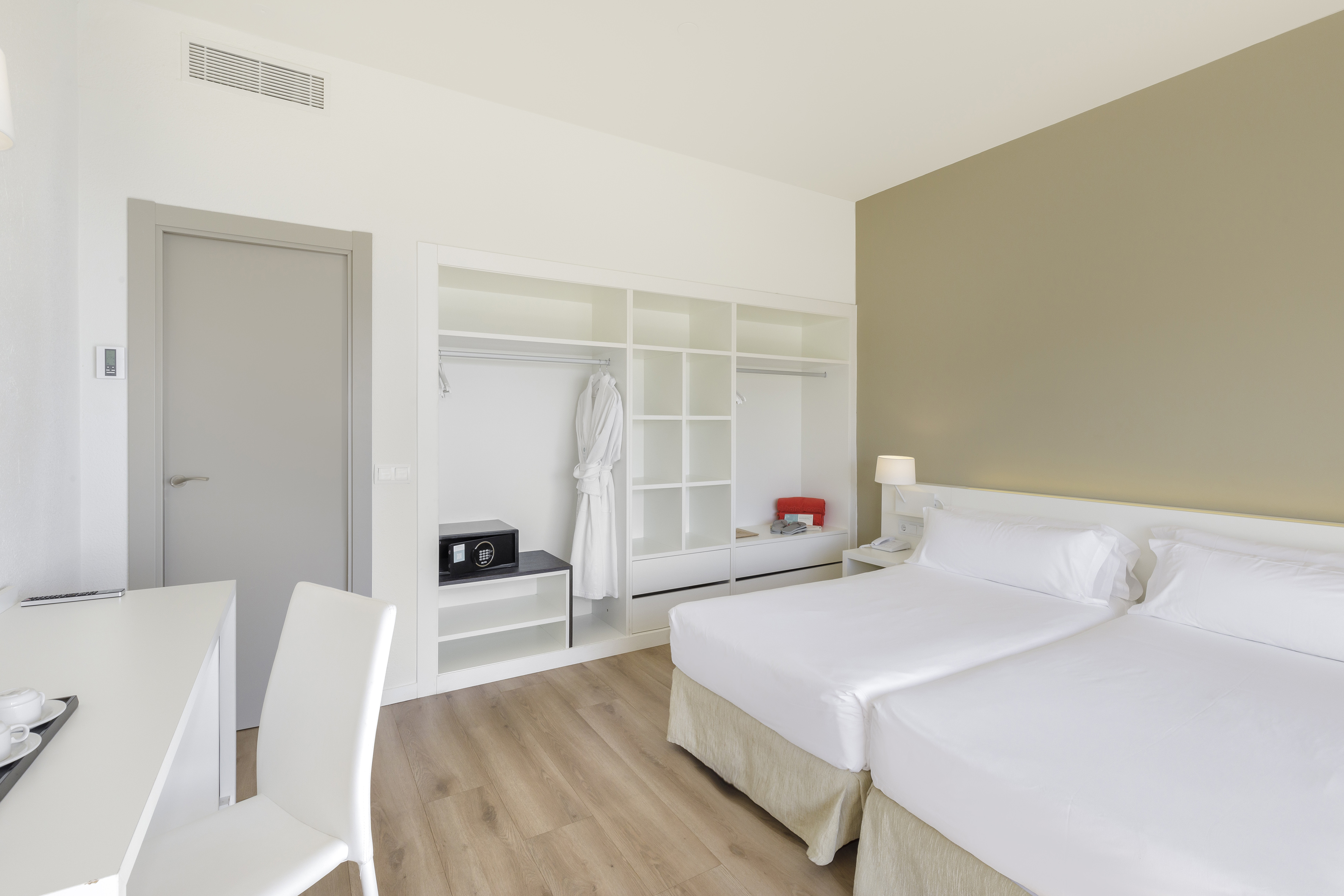 Modern hotel room with twin beds, white wardrobe, safe, and wooden floor.