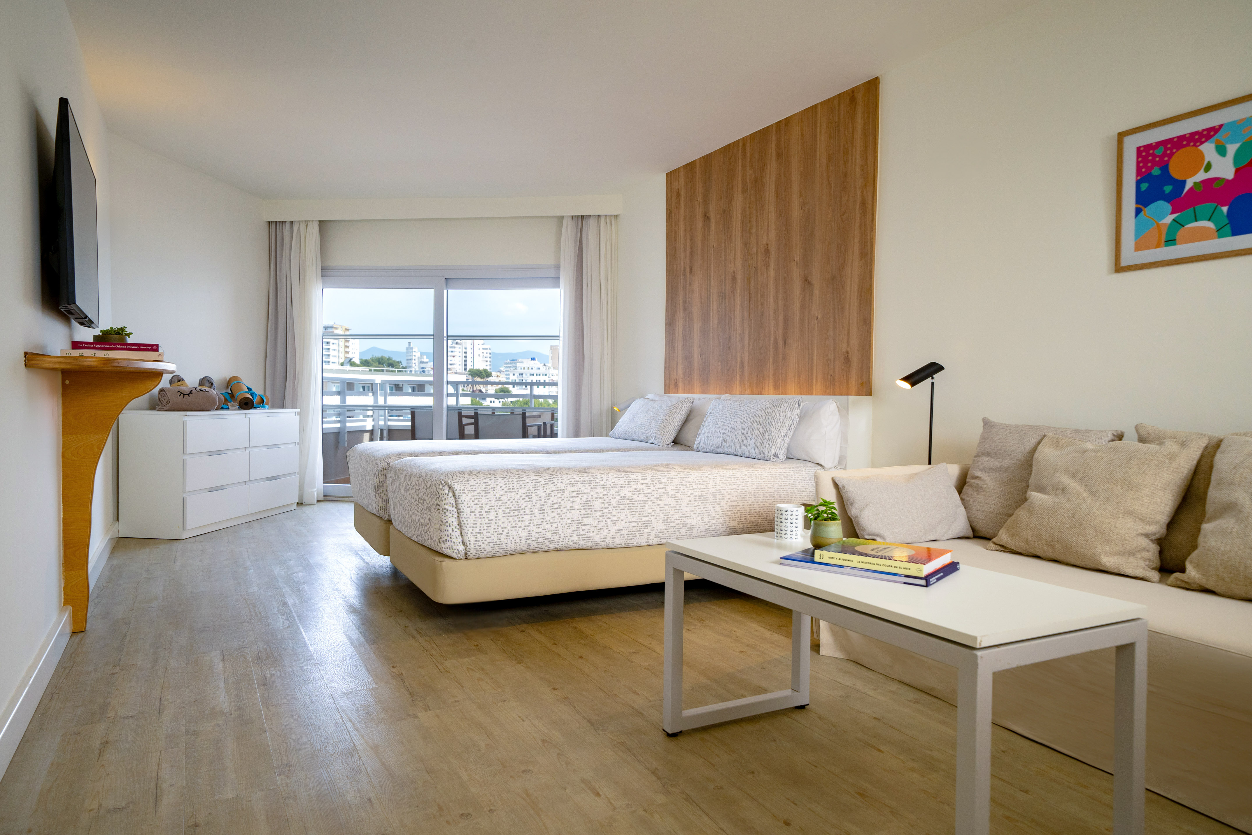 Modern hotel room with twin beds, sofa, balcony city view, wood details.