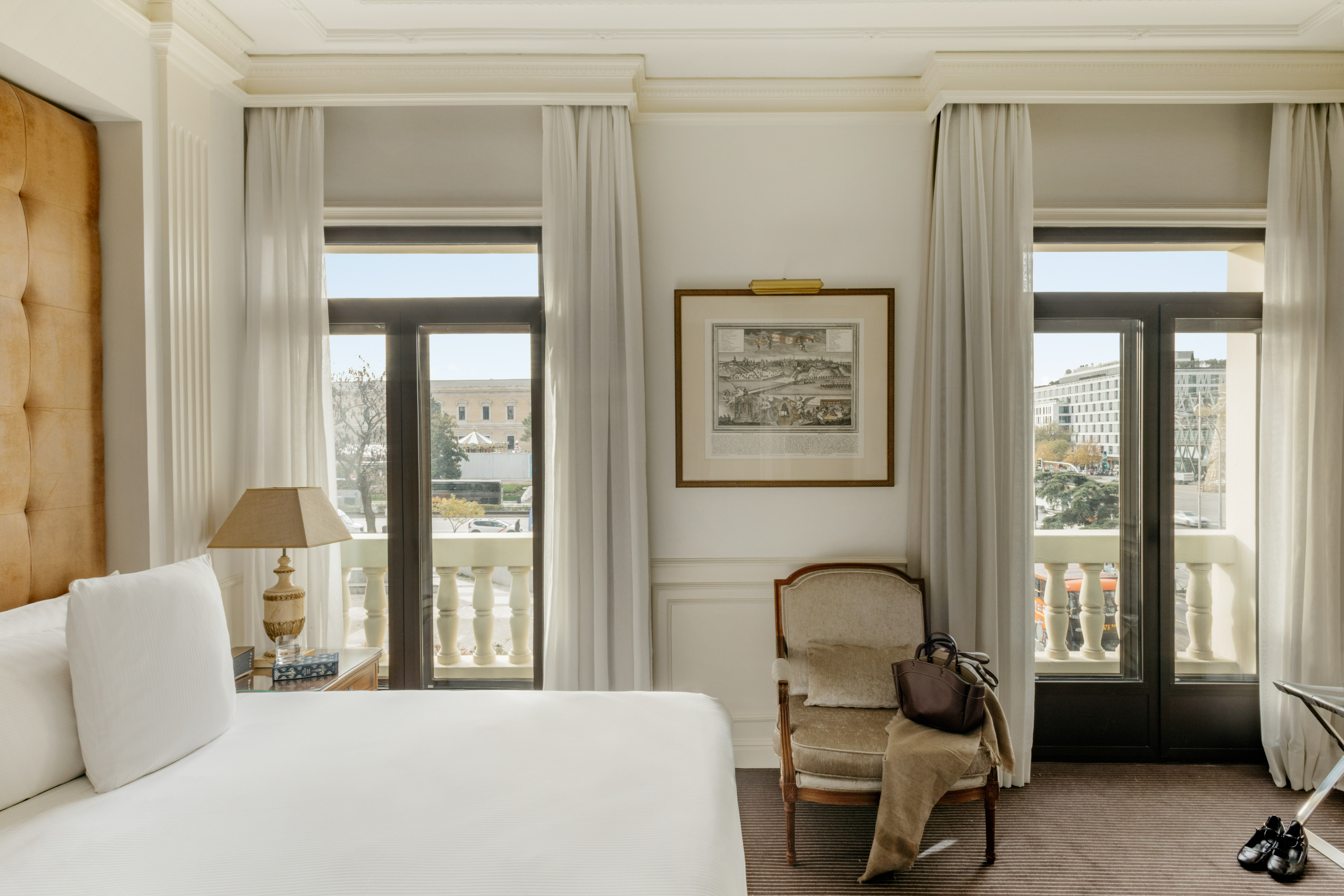 Elegant hotel room with white bed, city view balcony, and classic armchair.