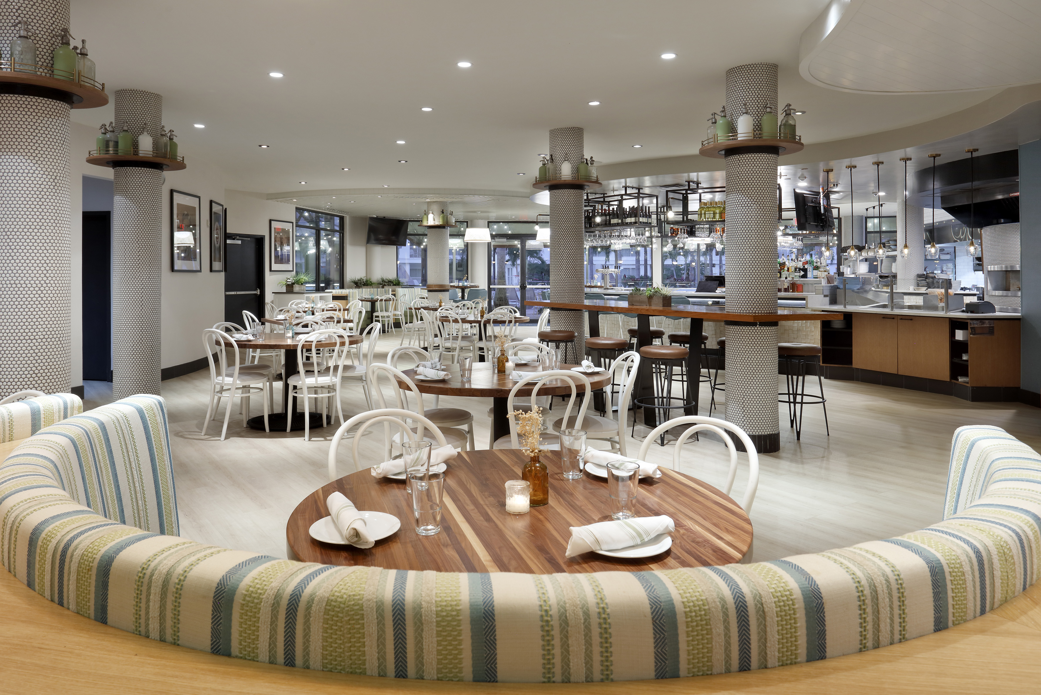 Bright restaurant interior, wood tables, white chairs, bar, and striped booth.