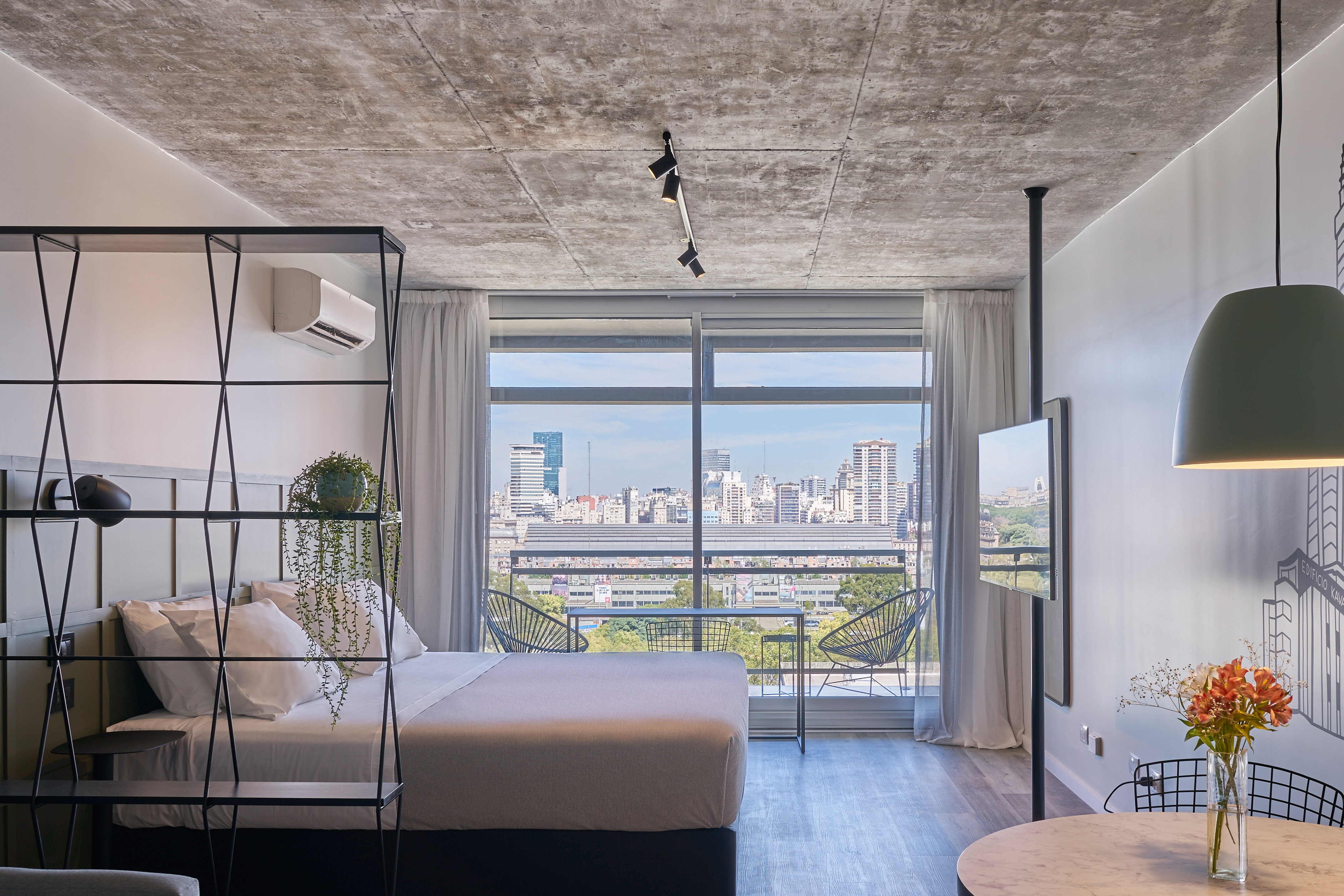 Modern hotel room with city skyline view, concrete ceiling, bed, and balcony.