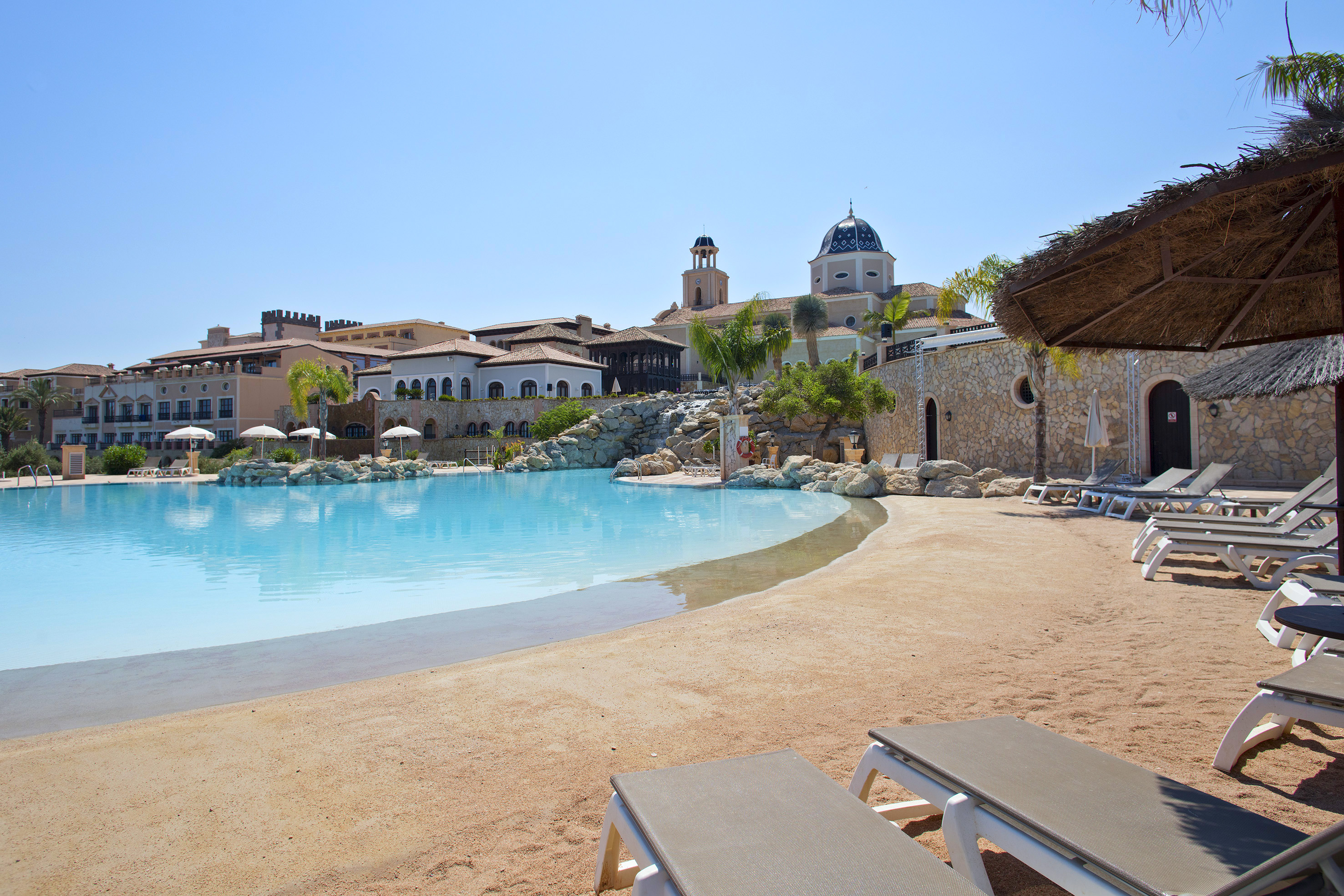 Resort lagoon pool with sandy beach, sun loungers, and Spanish-style buildings.