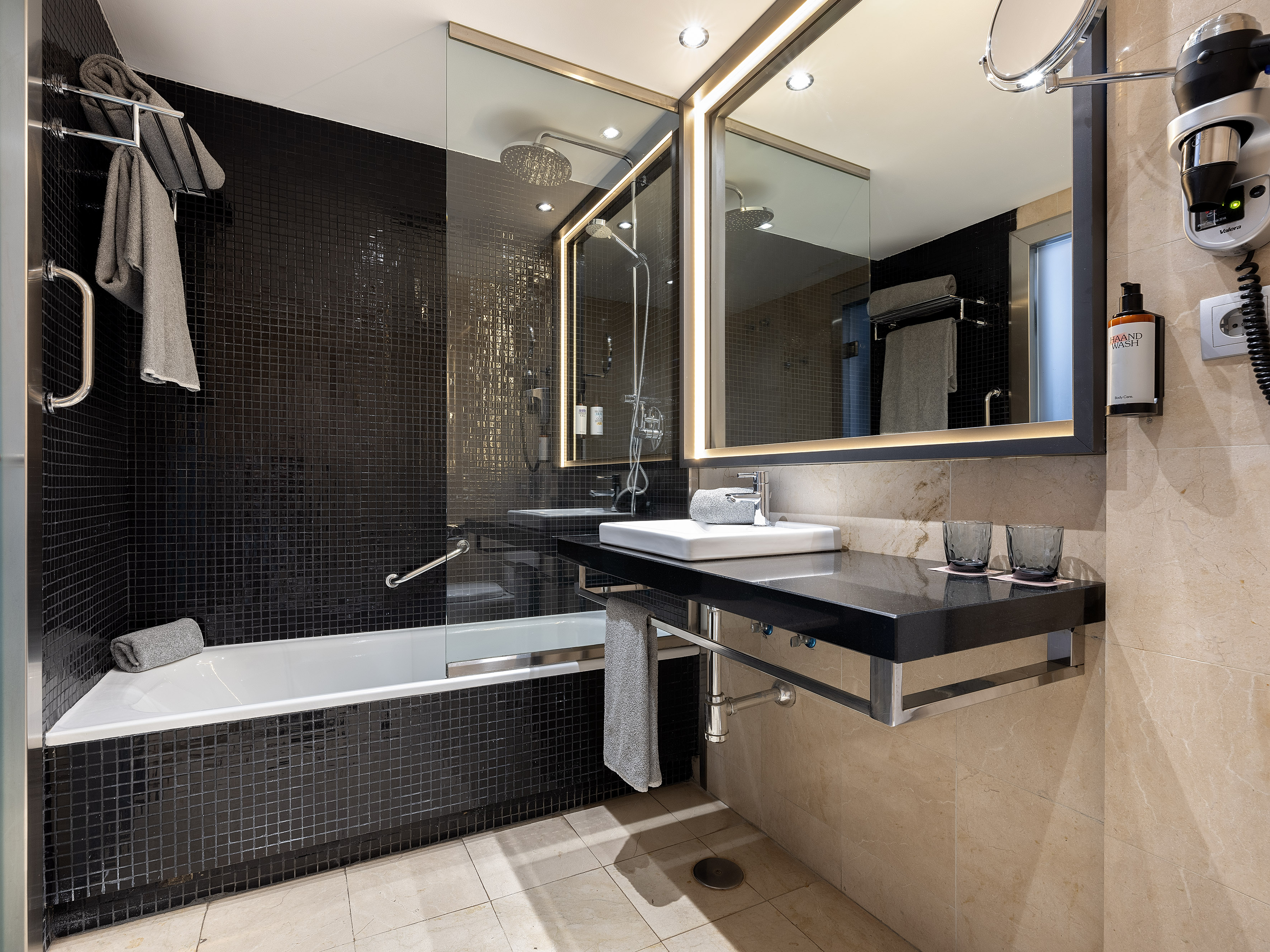 Modern hotel bathroom with black mosaic tiles, bathtub, shower, and lighted vanity.