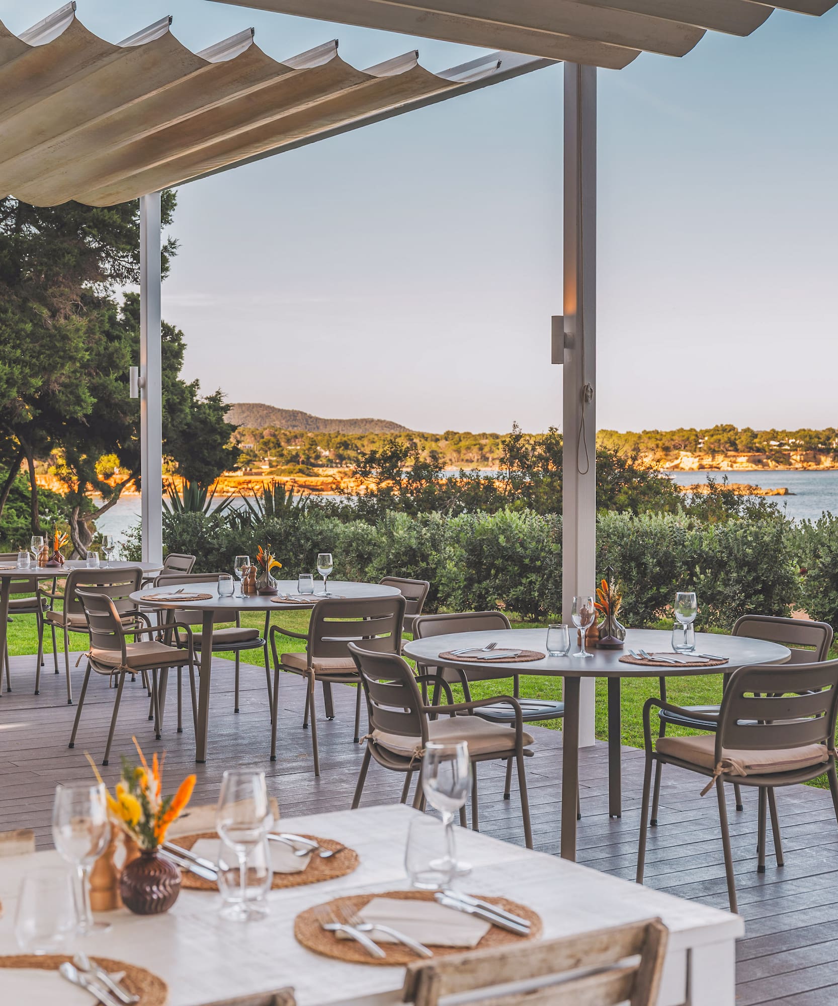 Outdoor restaurant terrace with sea view, set tables under an awning, lush green landscape.
