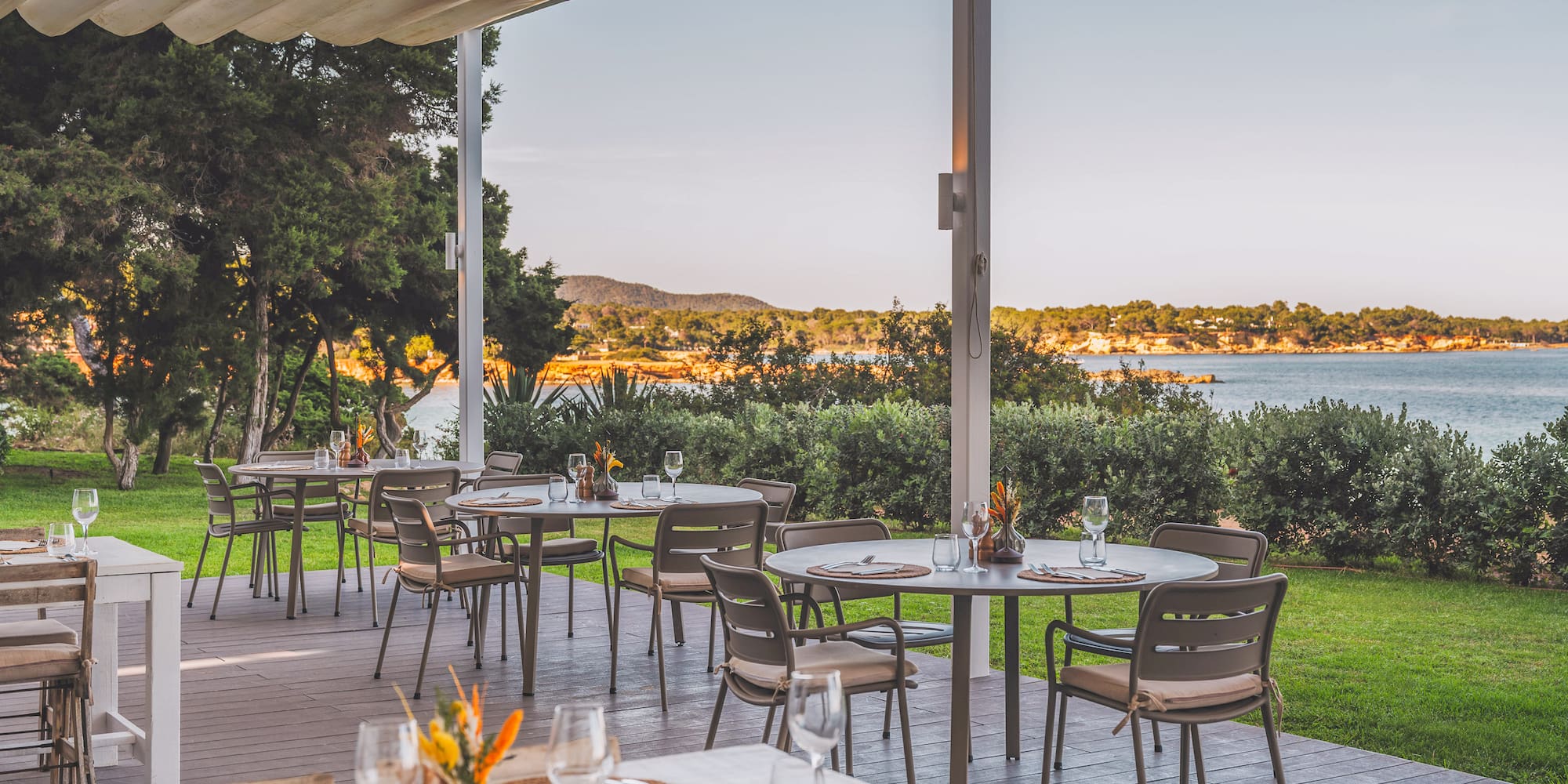 Outdoor restaurant terrace with sea view, set tables under an awning, lush green landscape.
