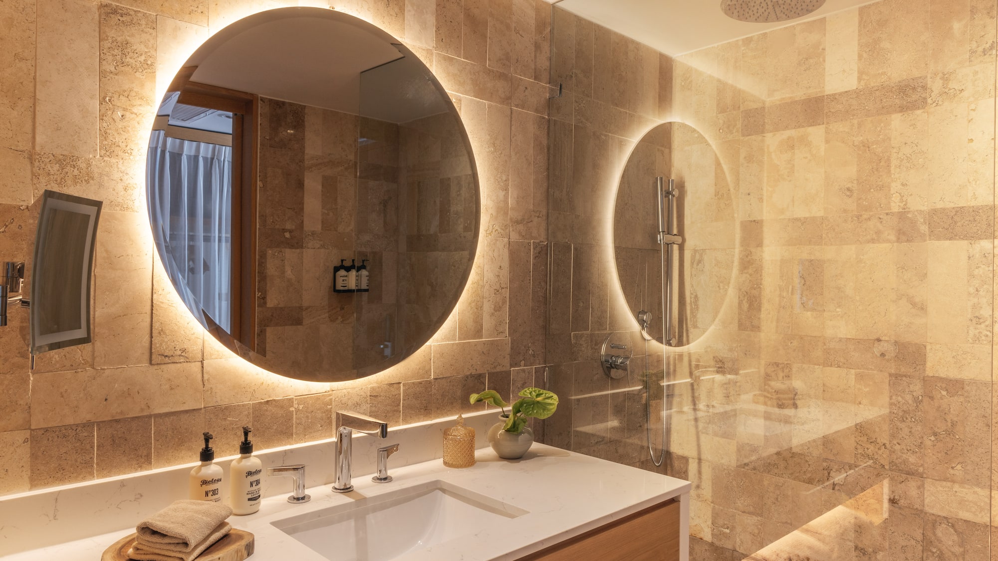 Modern bathroom with backlit mirror, white vanity, and stone-tiled shower.