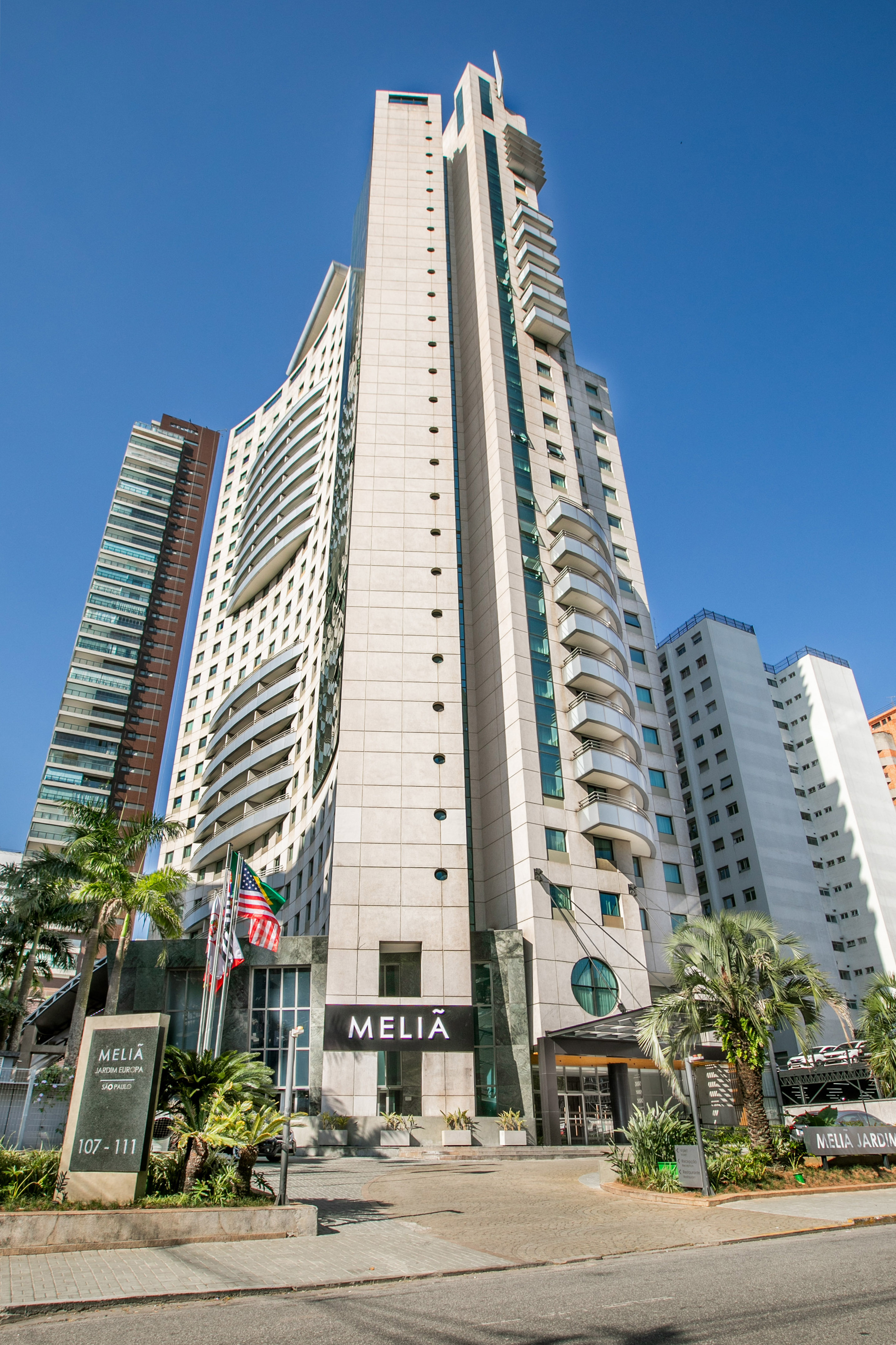 Tall Melia hotel building and modern skyscrapers under blue sky with palm trees.