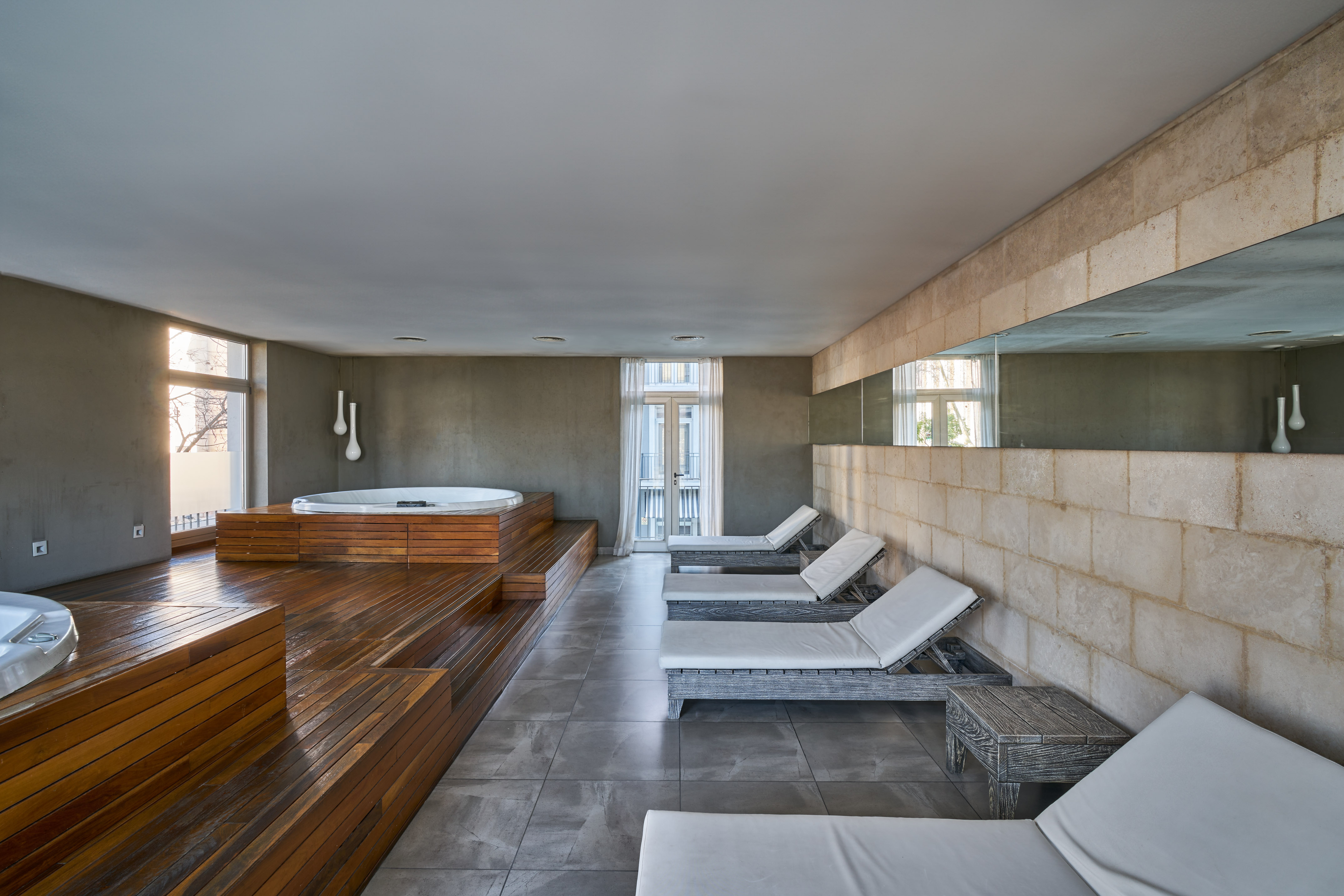 Modern spa interior with jacuzzis, wooden deck, lounge chairs, and stone wall.