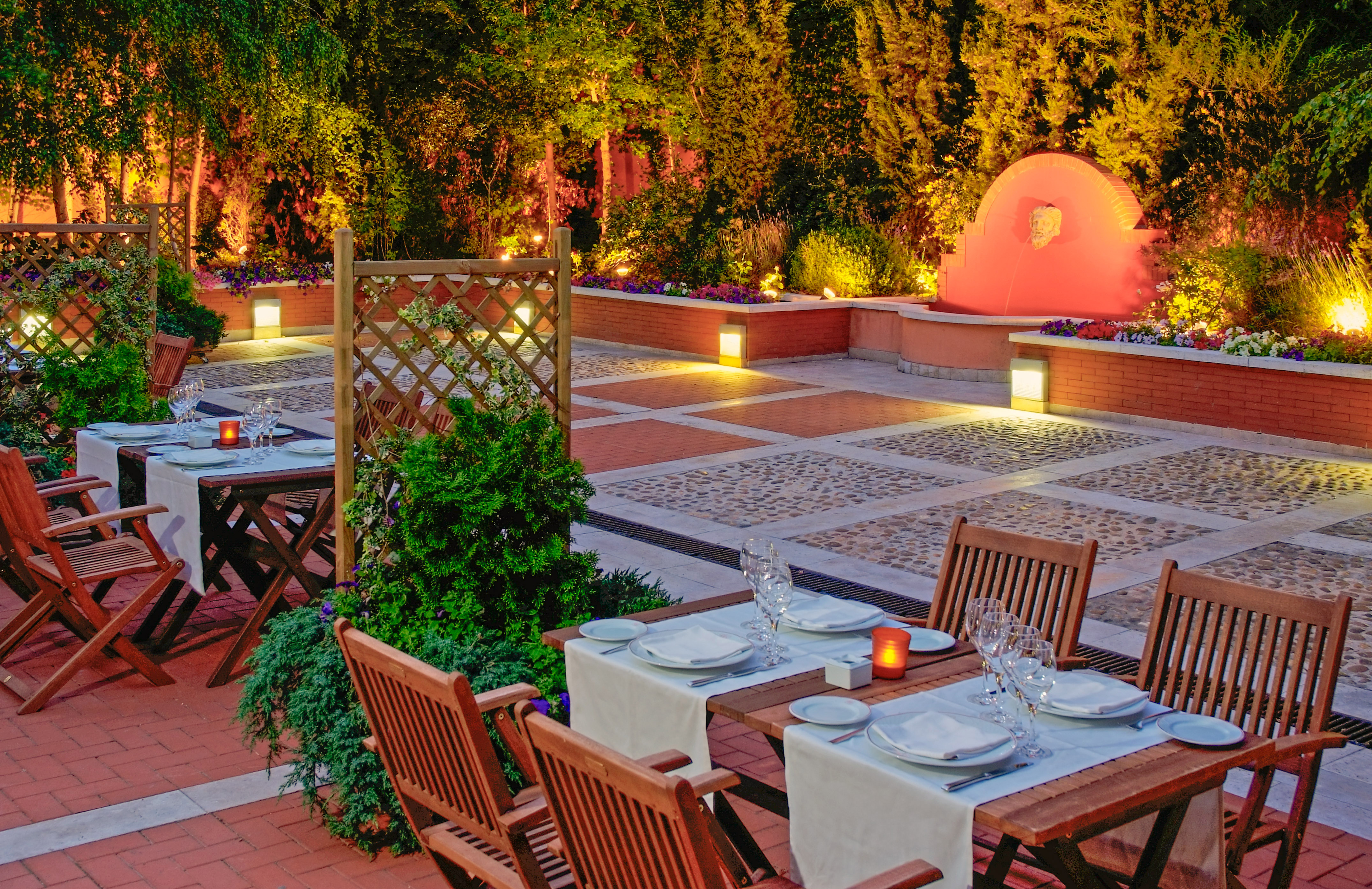 Terraza de restaurante exterior con mesas, árboles iluminados y fuente roja. - Meliá Recoletos, Valladolid