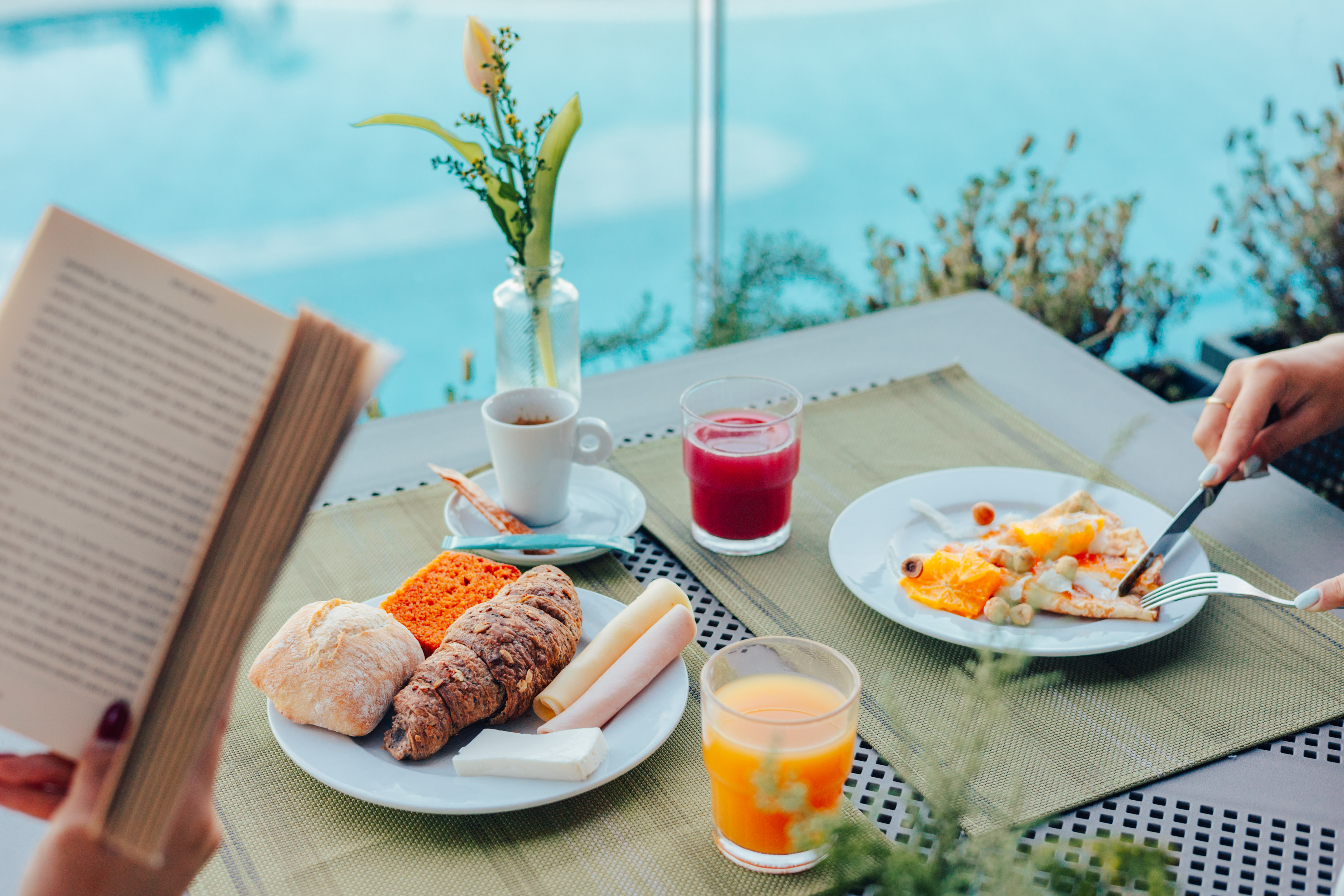 Outdoor breakfast by pool: pastries, coffee, juices, and person reading.