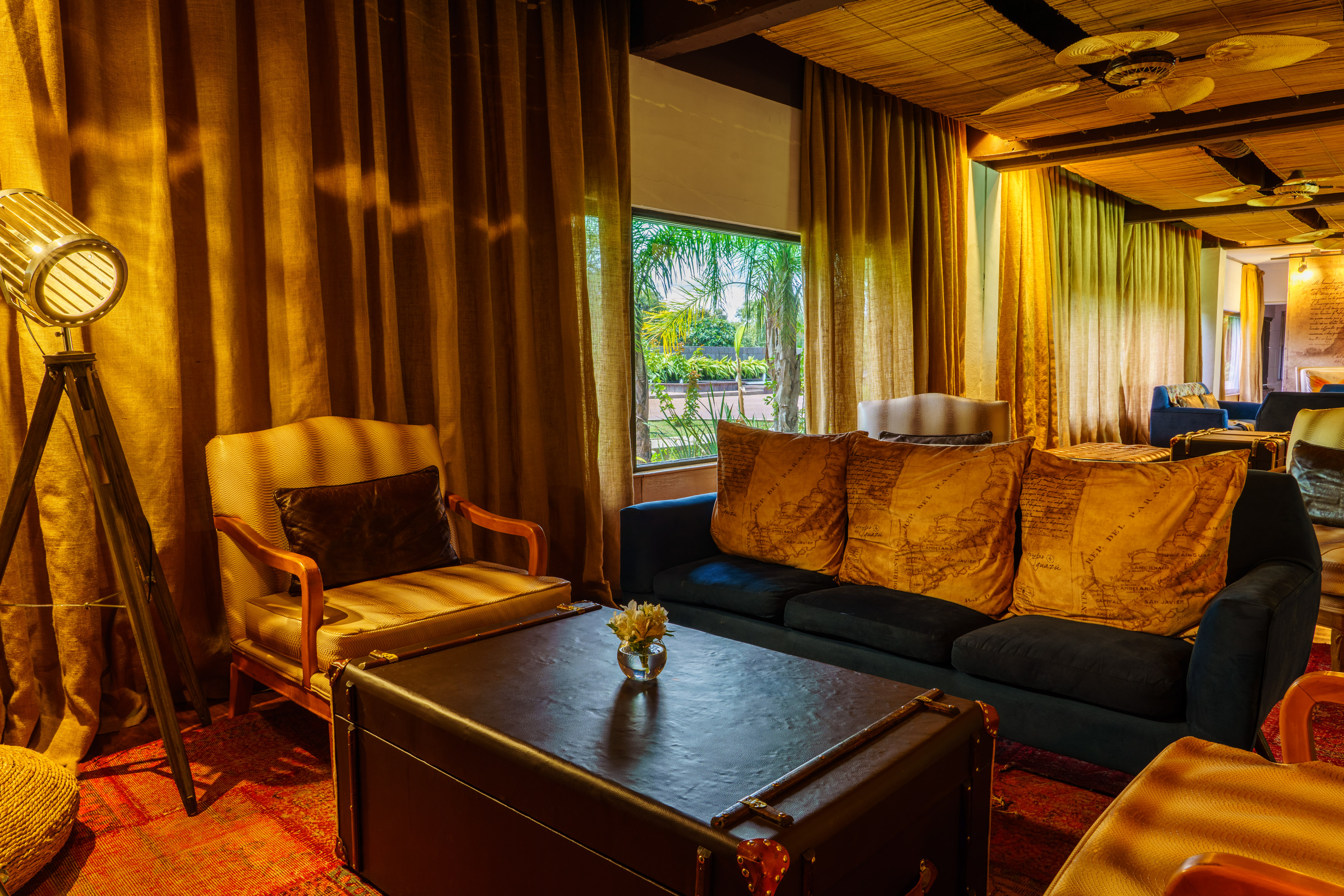 Warm tropical lounge with blue sofa, gold pillows, armchair, and lush window.