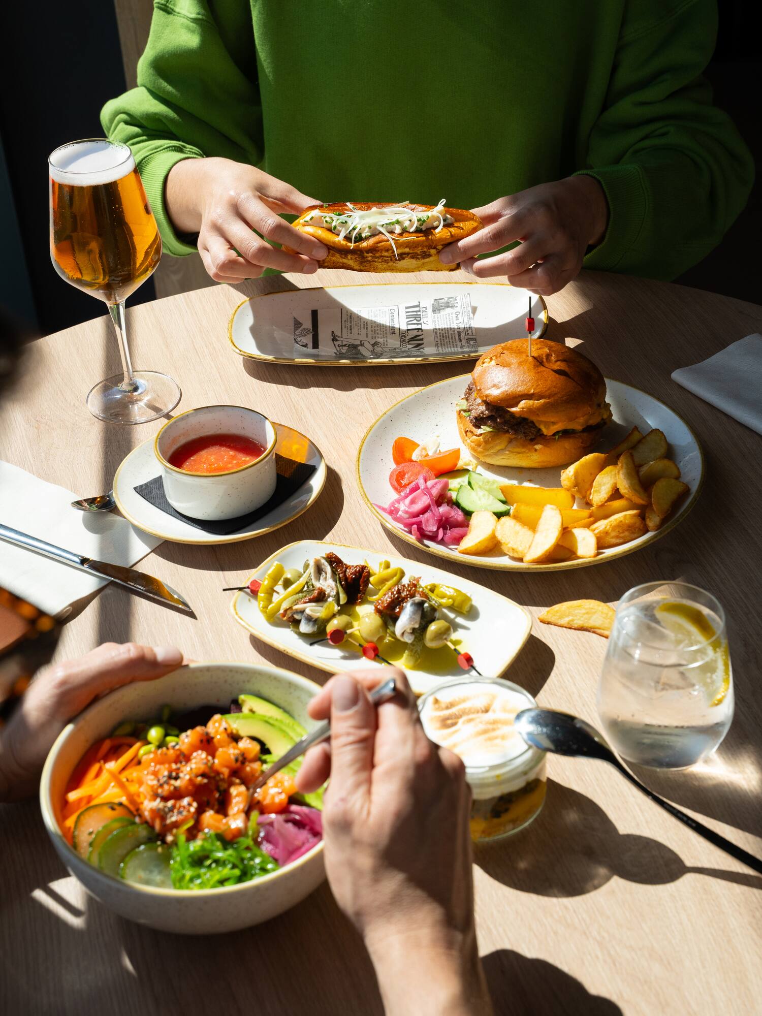Restaurant meal with burger, hot dog, poke bowl, fries, beer in sunlight.