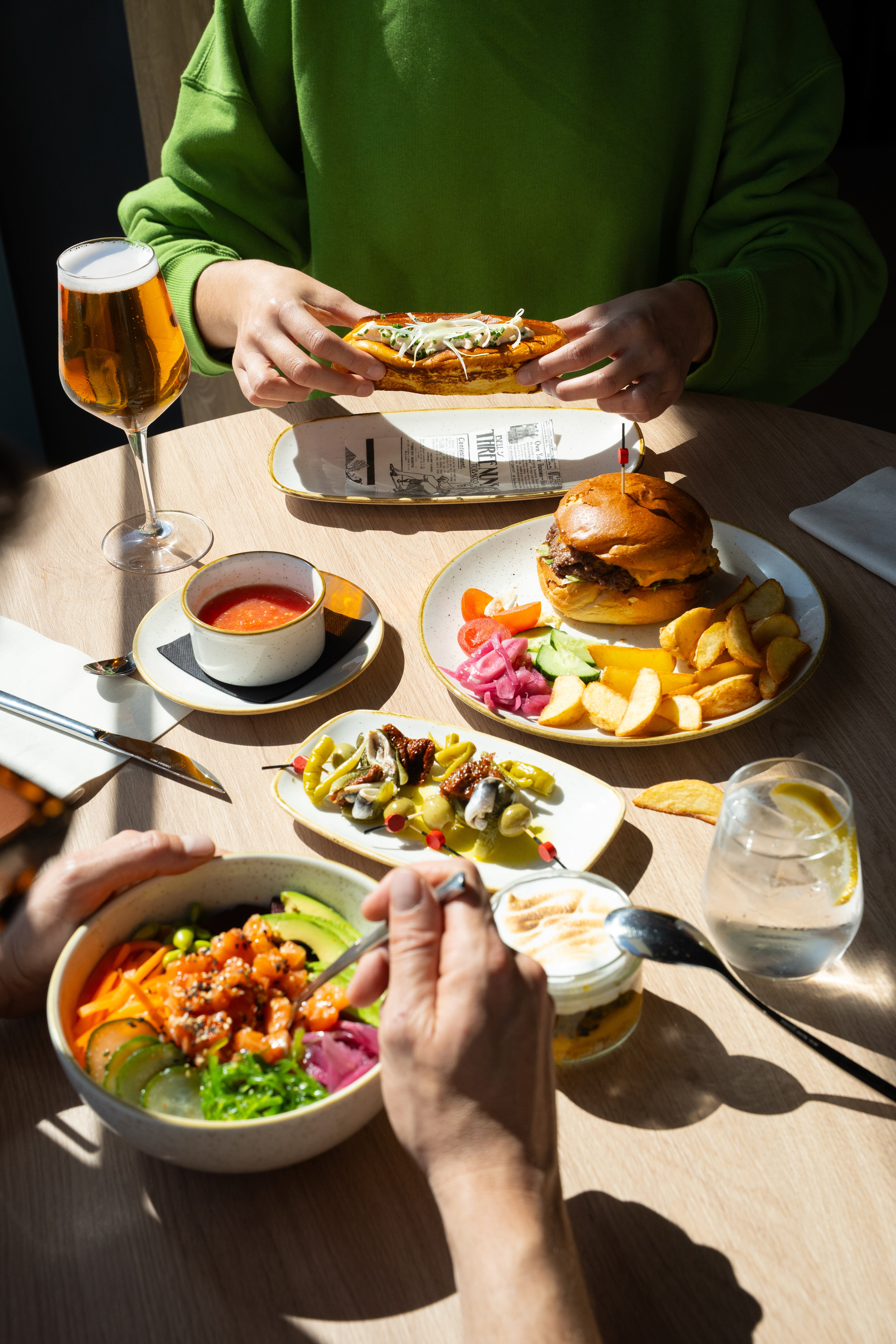 Restaurant meal with burger, hot dog, poke bowl, fries, beer in sunlight.