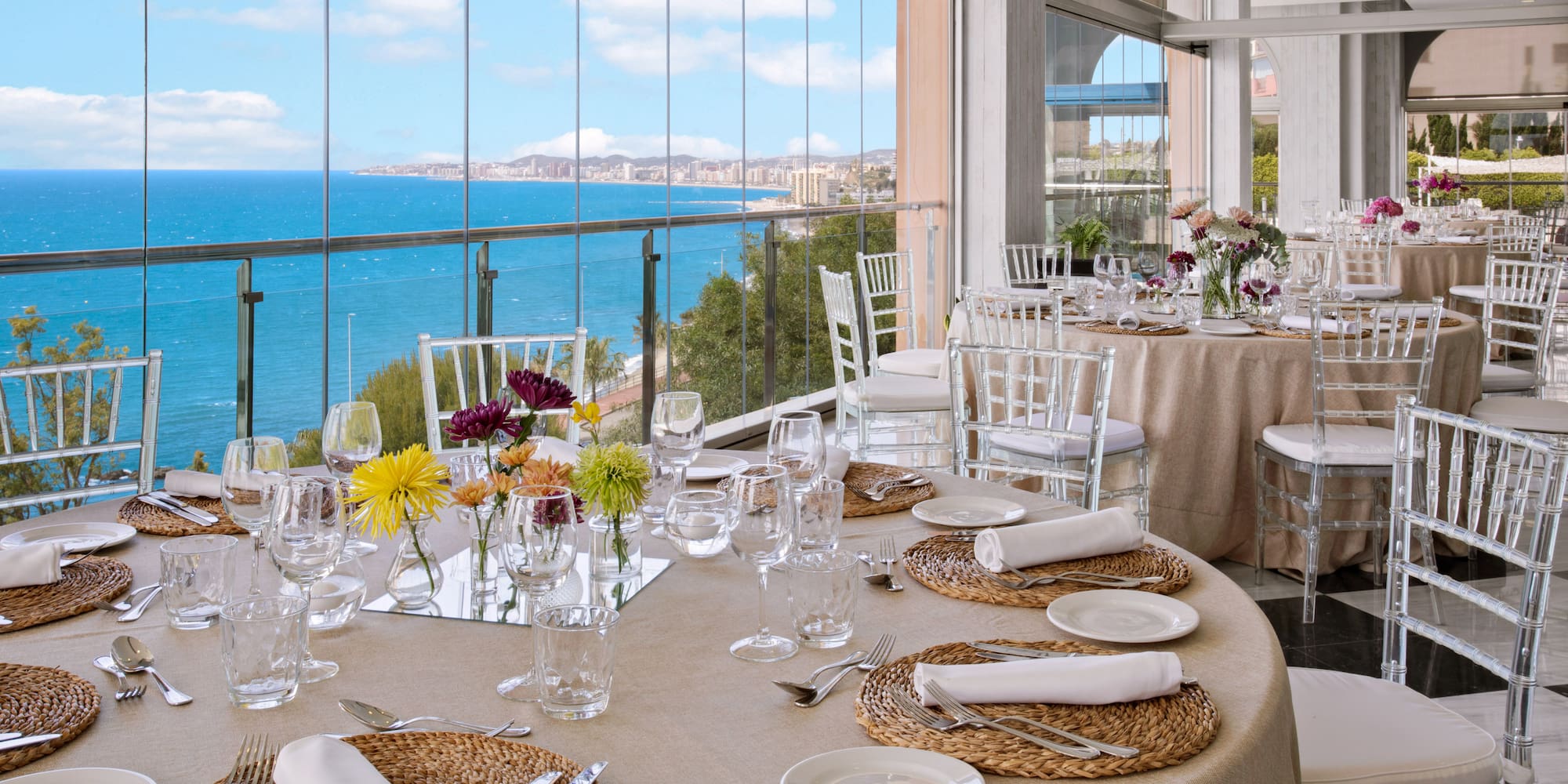 Elegant banquet tables, floral decor, panoramic sea and coastal city view.