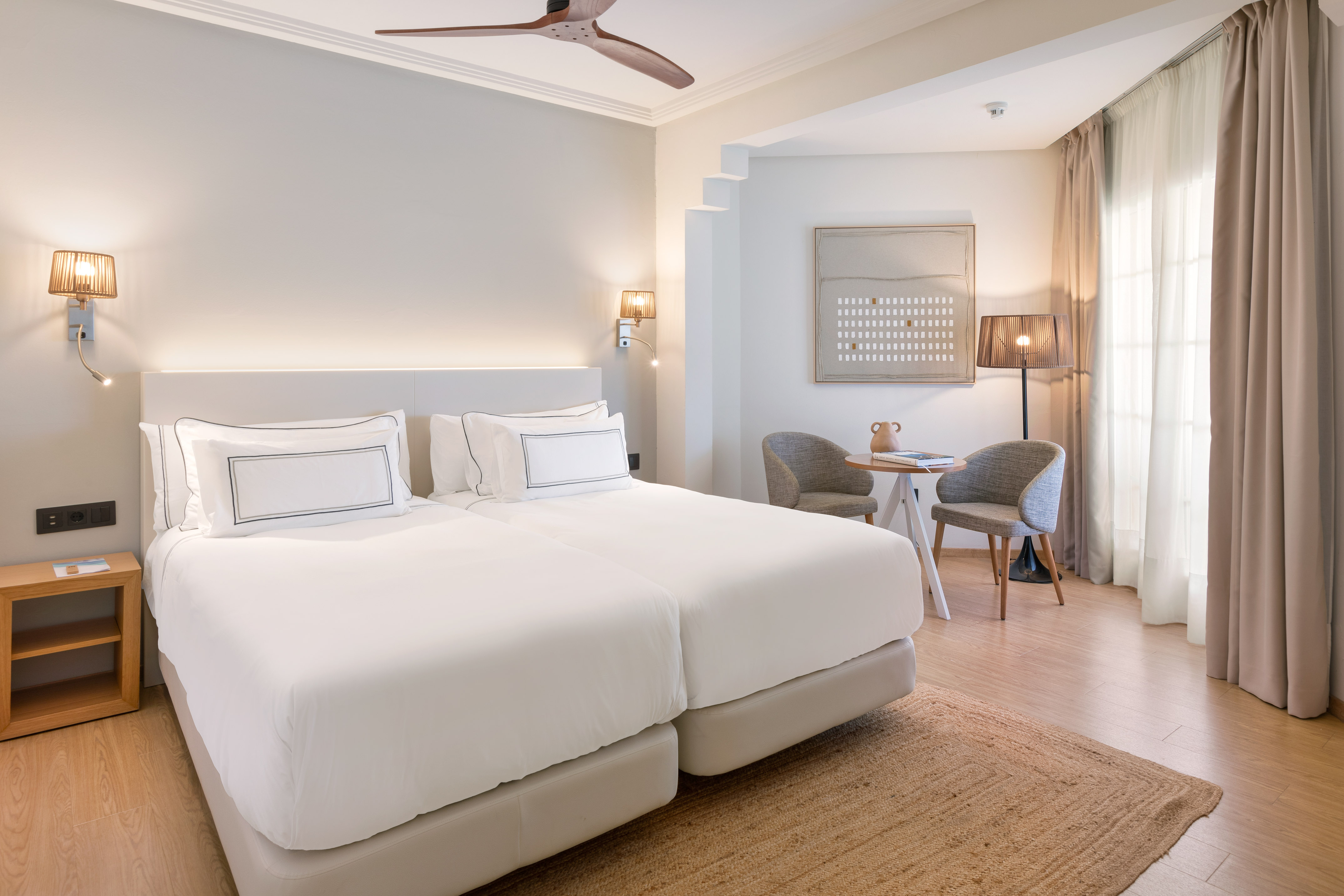 Modern hotel room with two single beds, neutral decor, and ceiling fan.