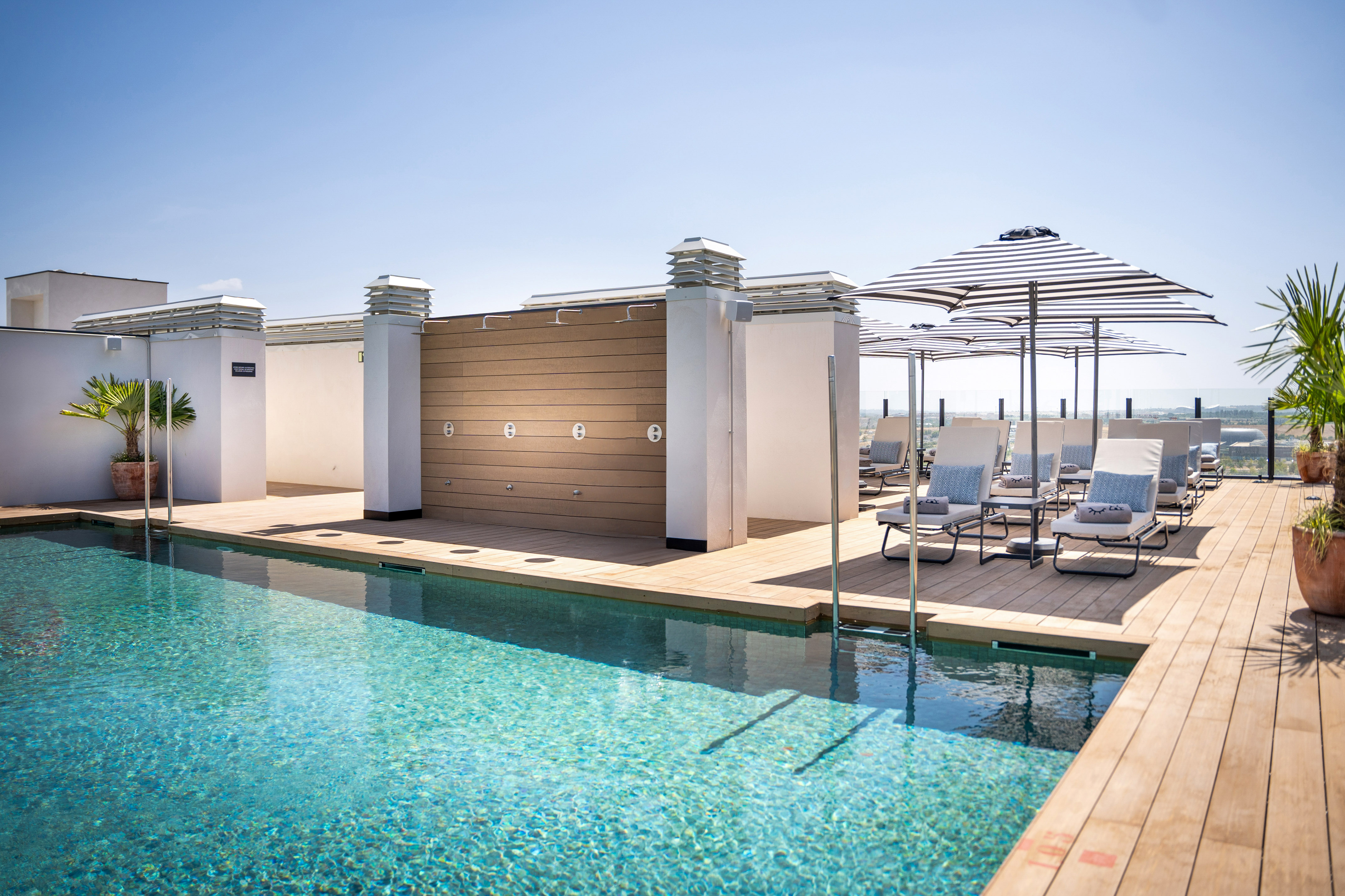 Rooftop swimming pool with wooden deck, lounge chairs, striped umbrellas, and showers.