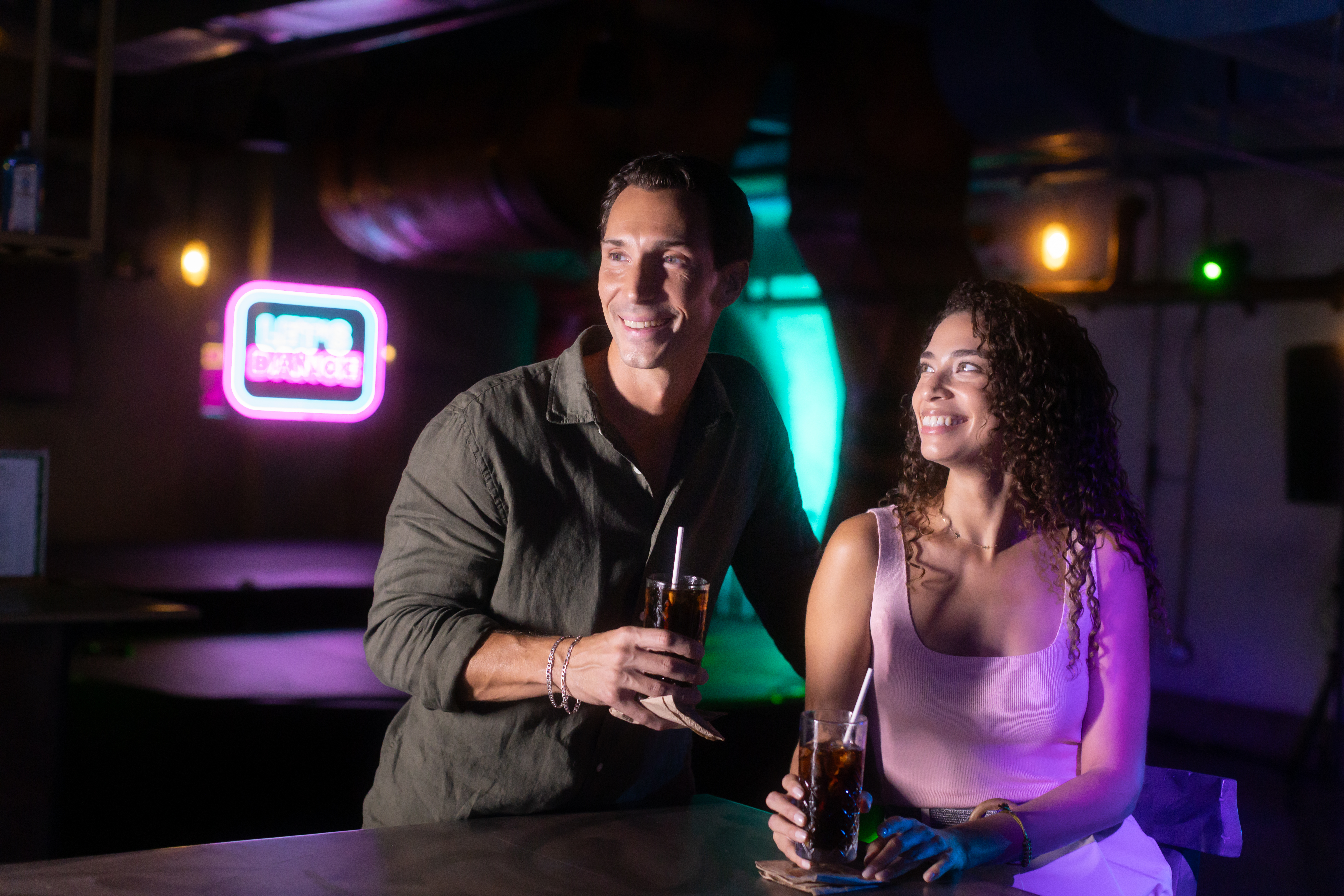 Happy couple smiling, holding drinks at a vibrant bar with neon lights.