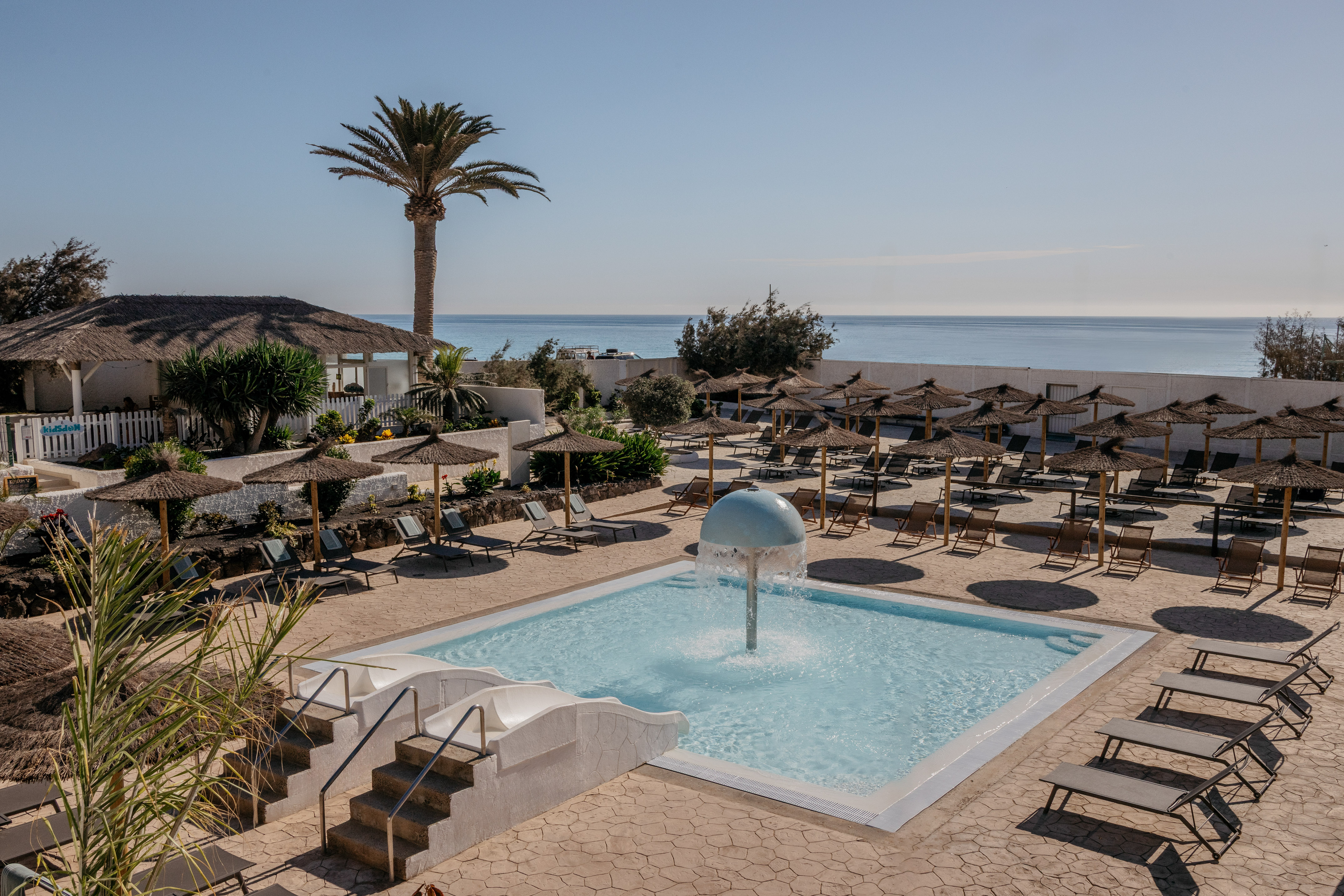 Hotel pool with fountain, sun loungers, palm tree, and ocean view.