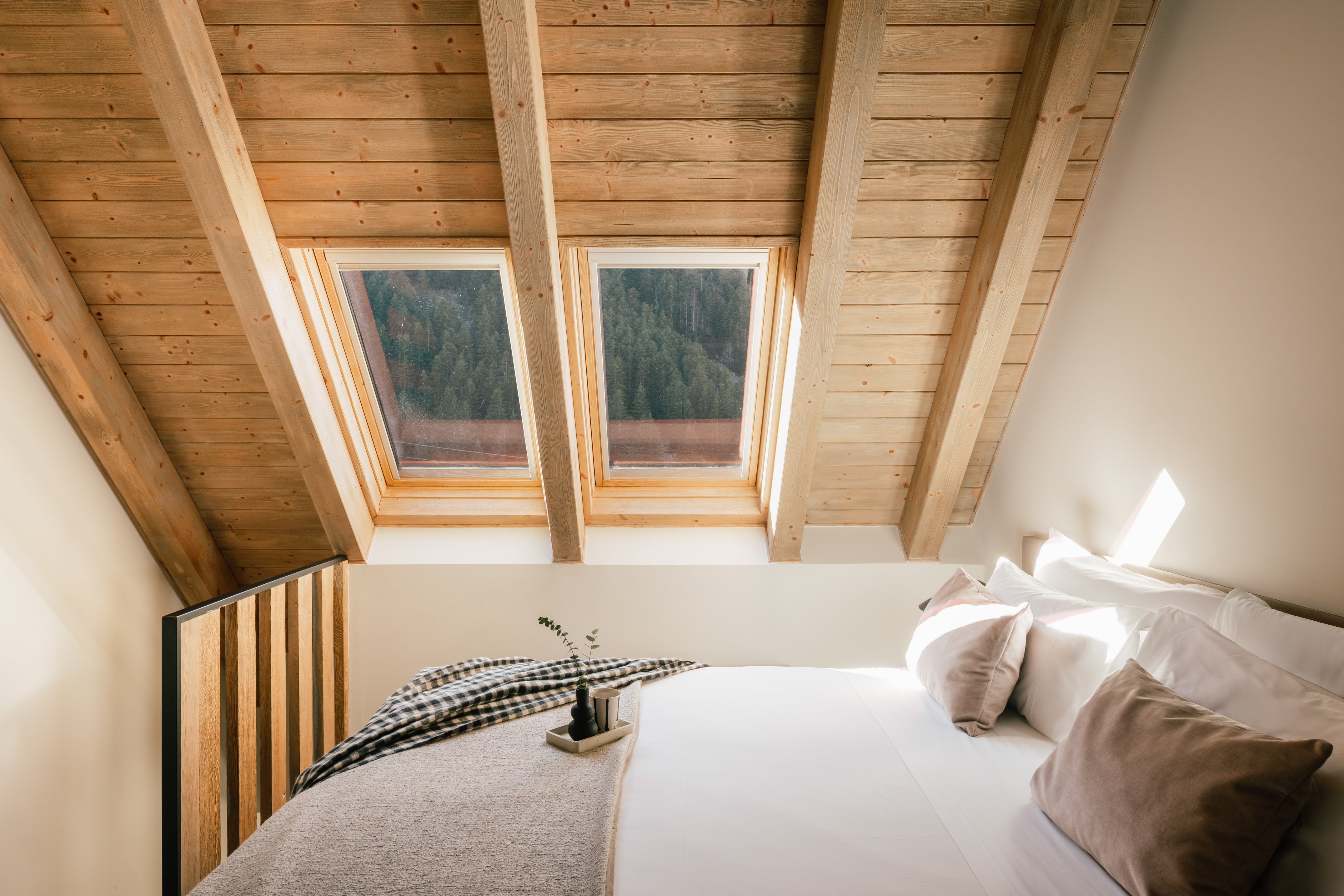 Cozy attic bedroom with wood ceiling, skylights, and forest view.