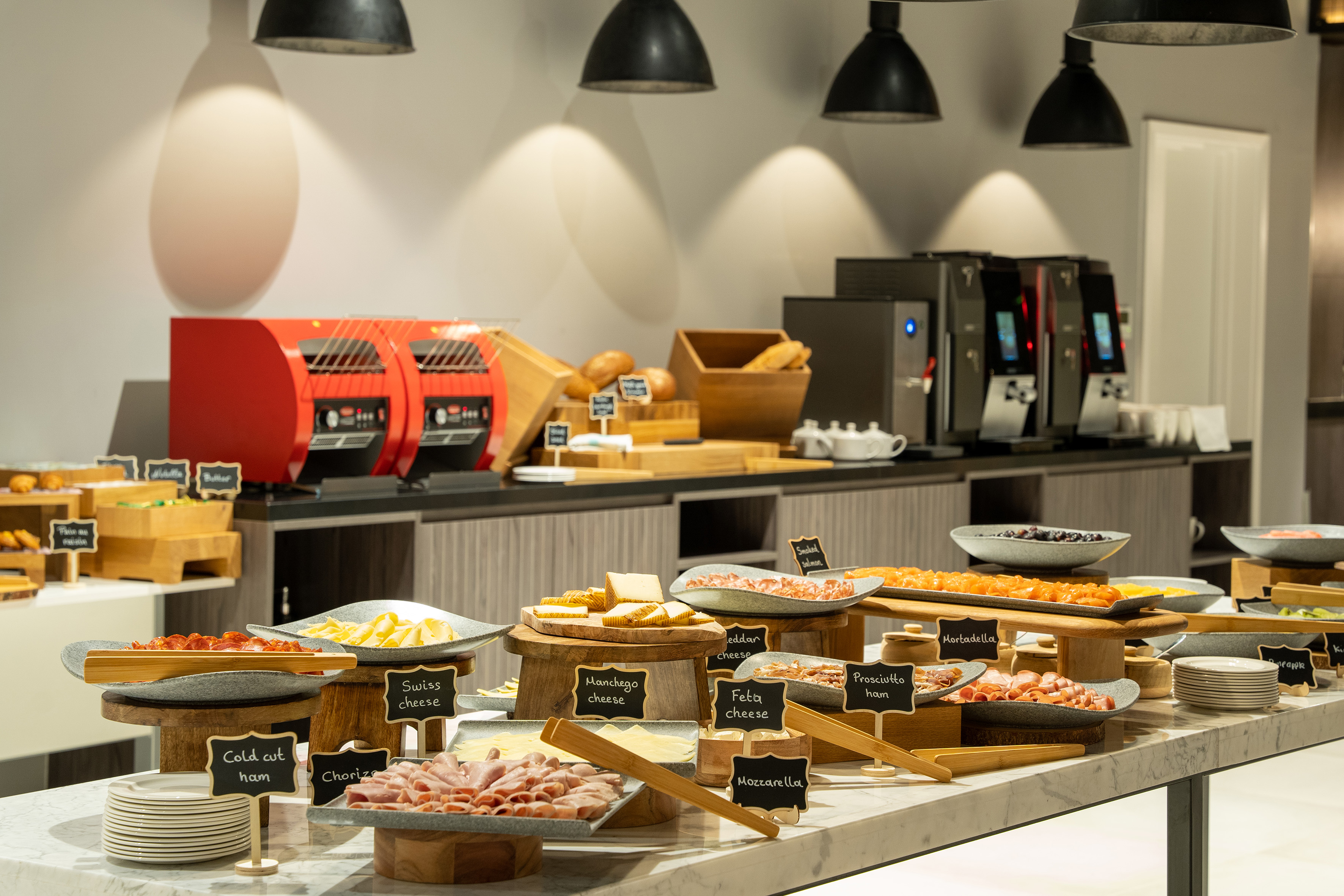 Breakfast buffet with various cold cuts, cheeses, bread, and coffee machines.