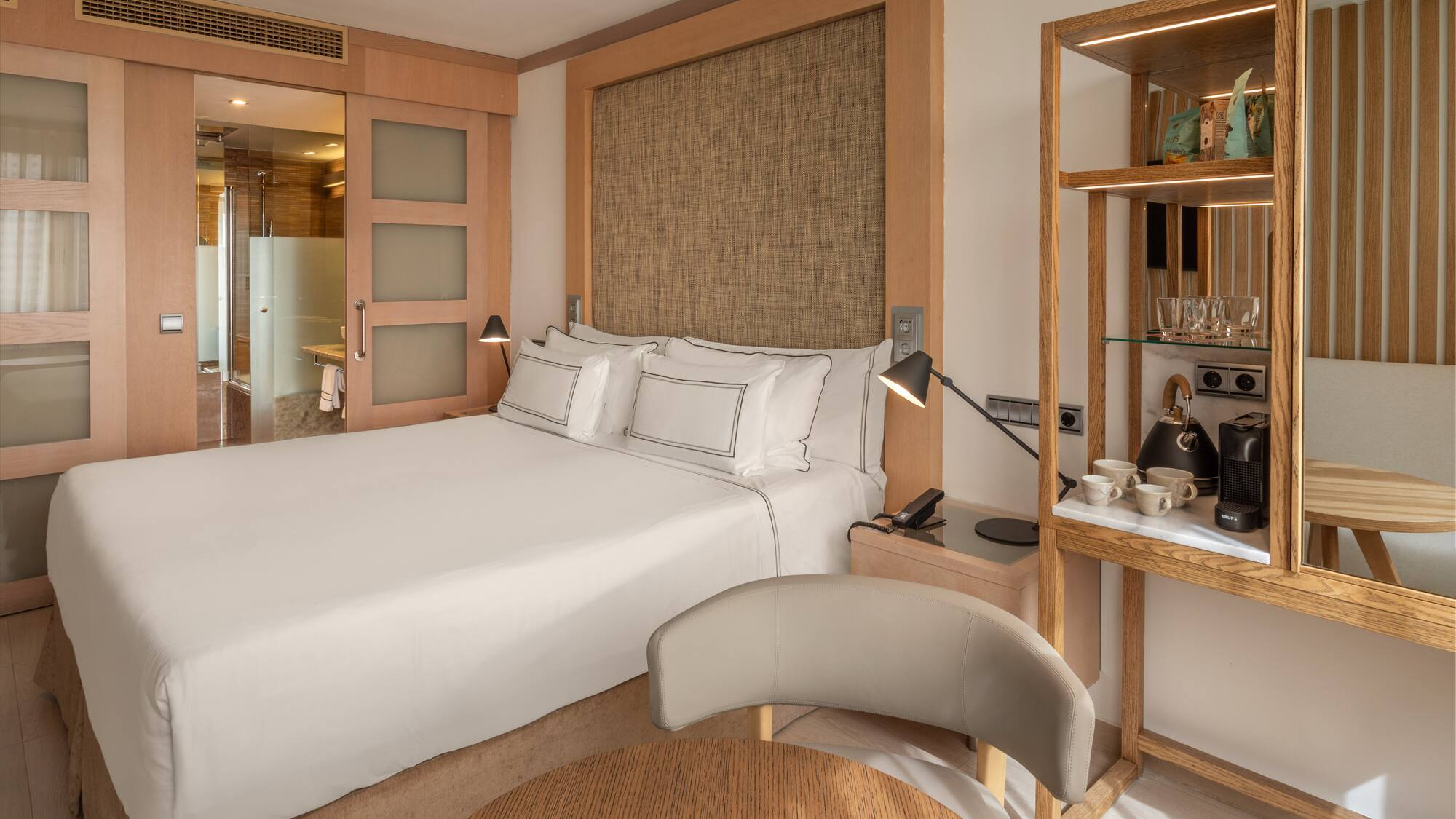 Modern hotel bedroom with king bed, wood accents, attached bath, and coffee maker.