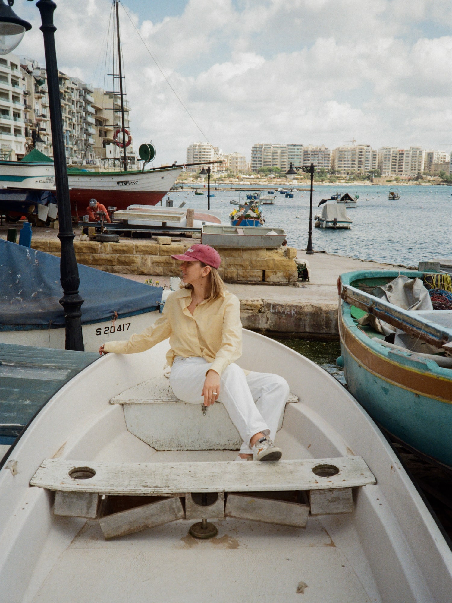 Woman in yellow shirt, red cap, sits in boat at a sunny Malta harbor.