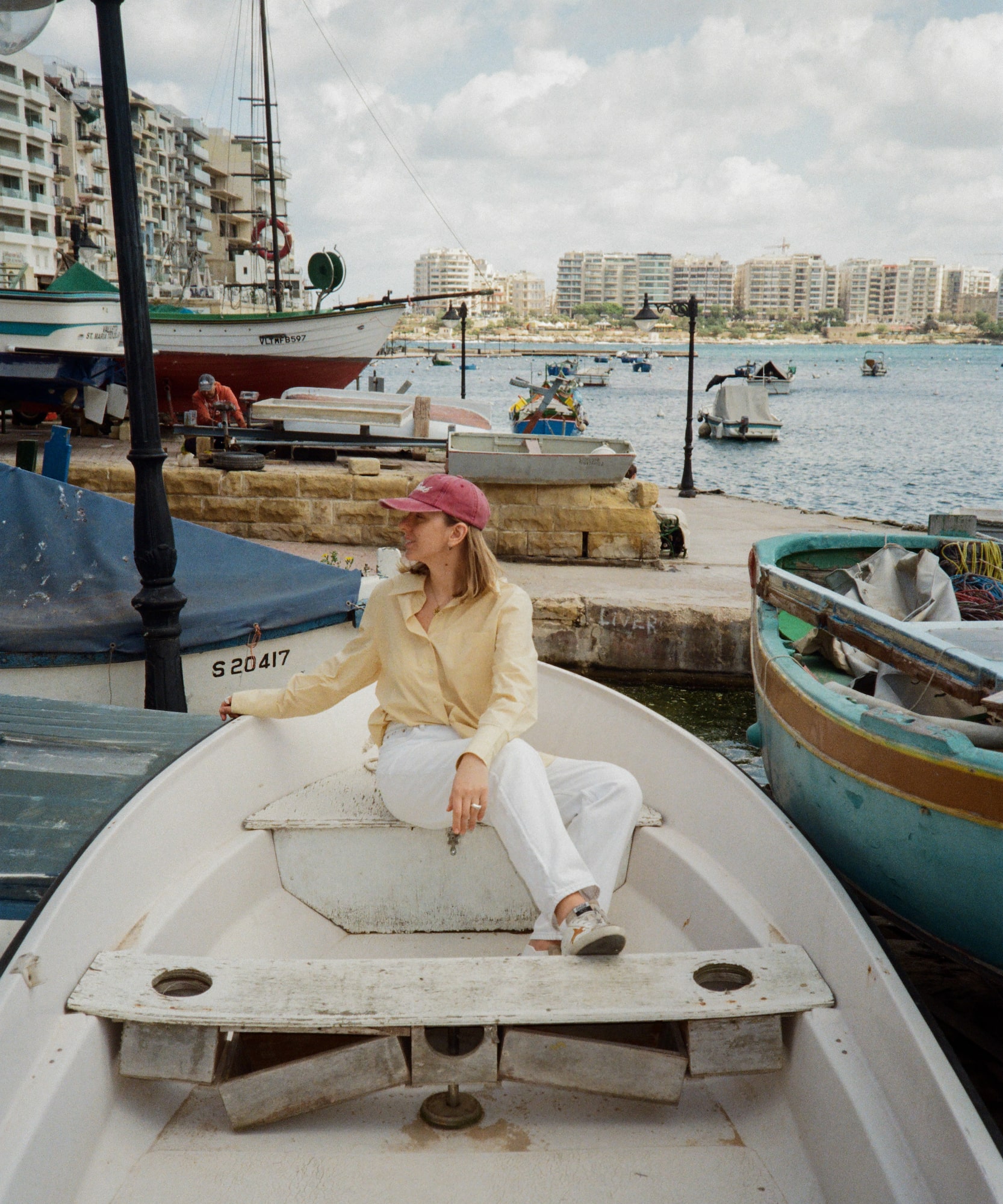 Woman in yellow shirt, red cap, sits in boat at a sunny Malta harbor.