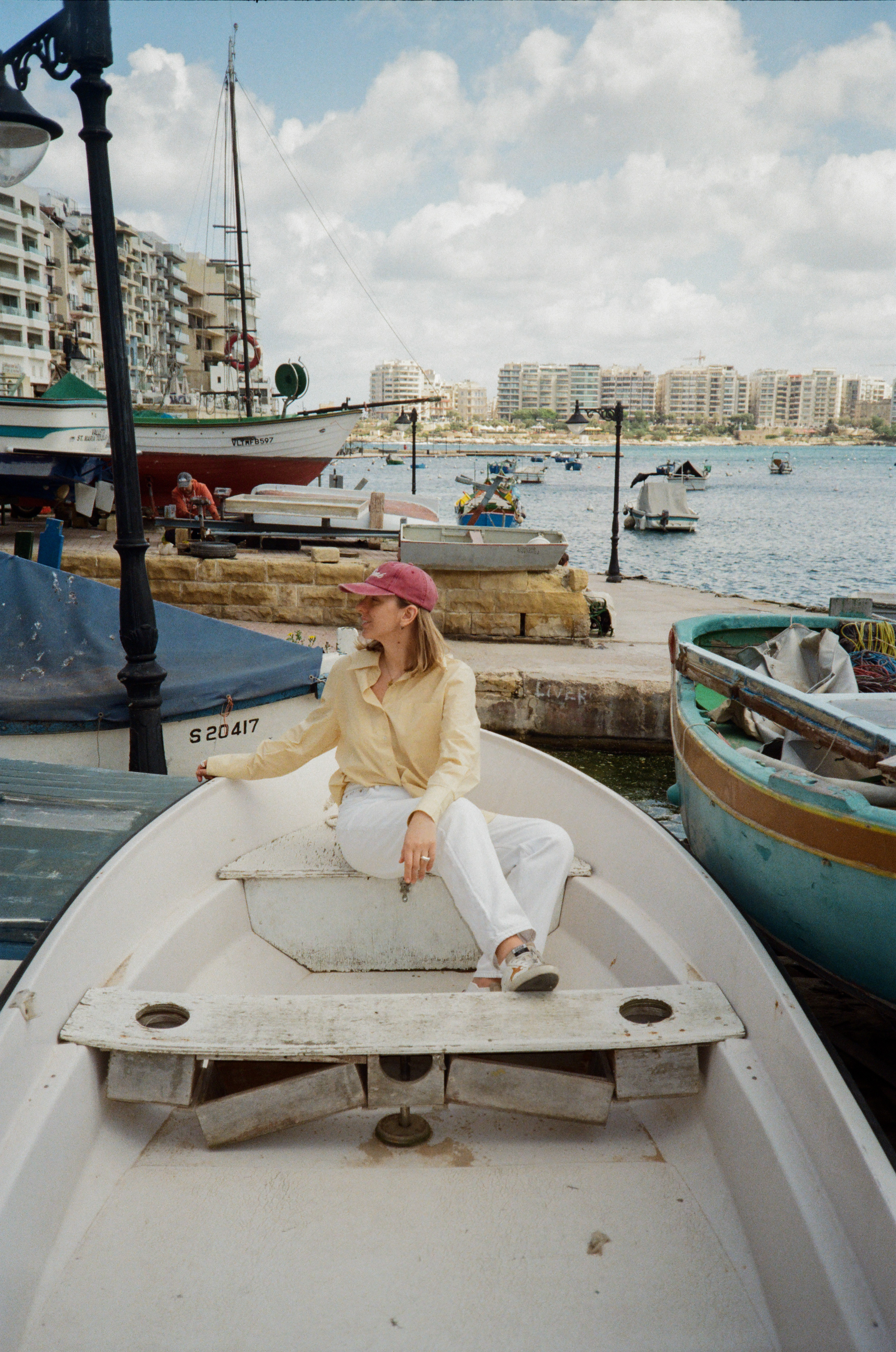 Woman in yellow shirt, red cap, sits in boat at a sunny Malta harbor.