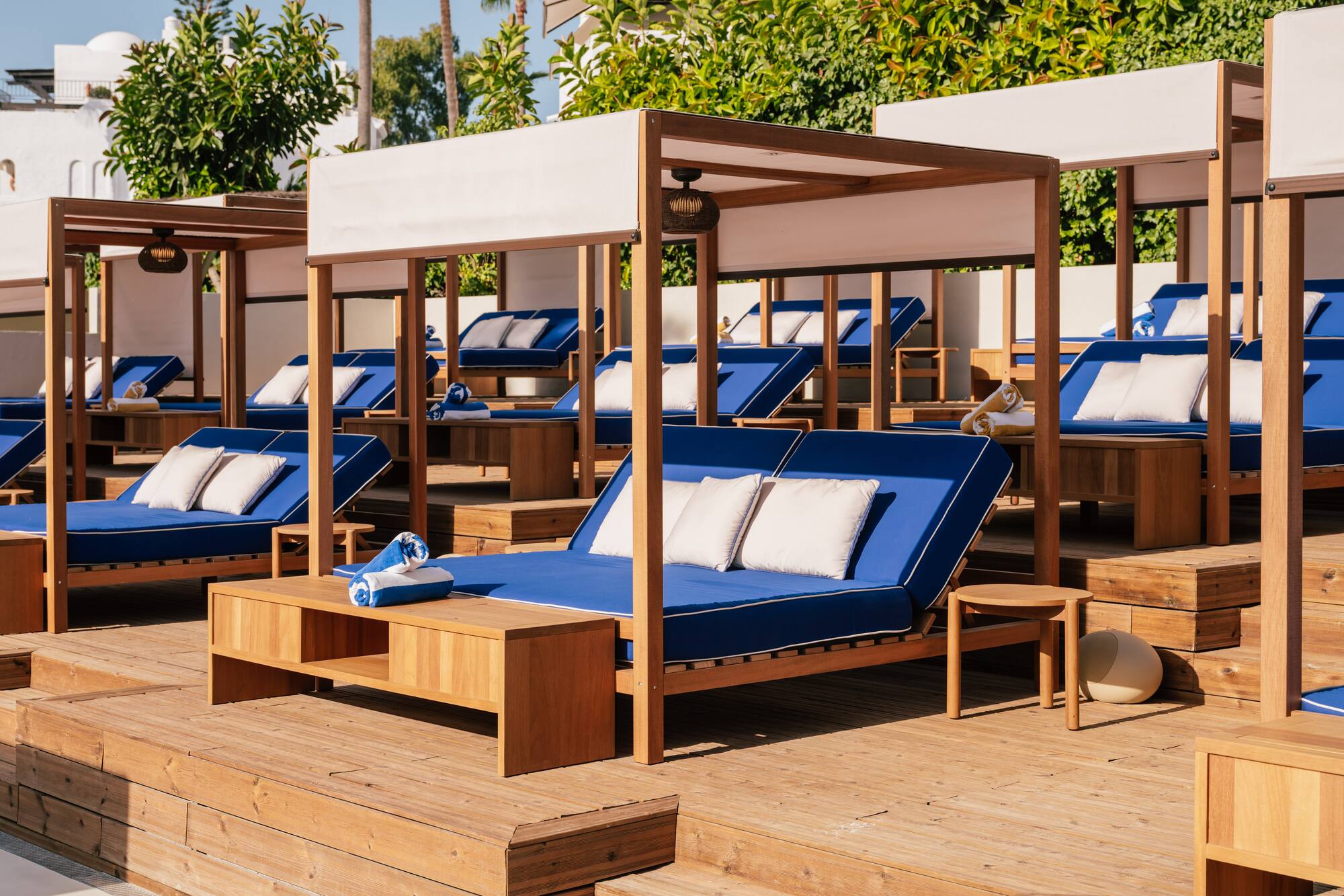Sunlit luxury resort cabanas with blue daybeds on a wooden deck.