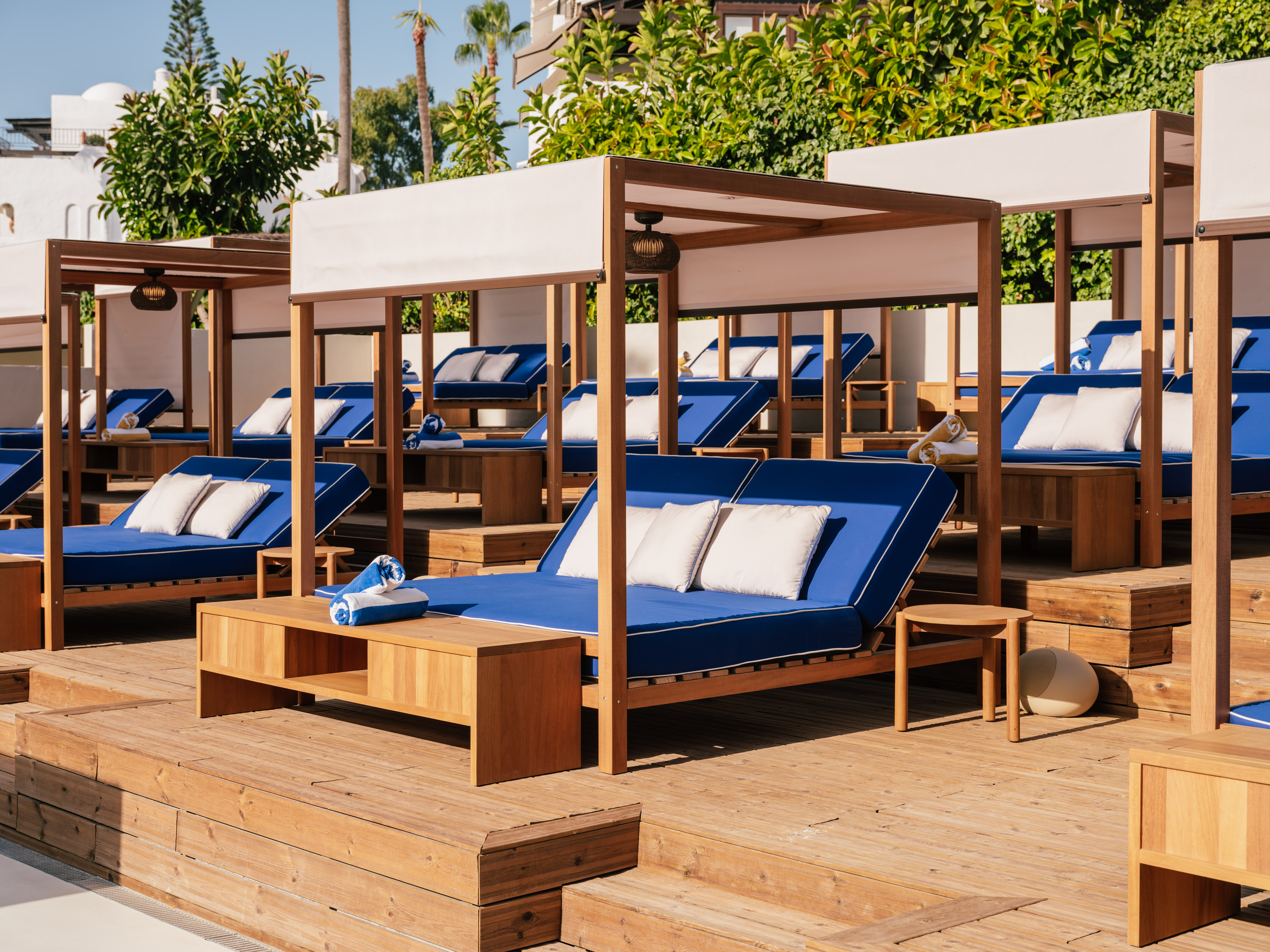 Sunlit luxury resort cabanas with blue daybeds on a wooden deck.