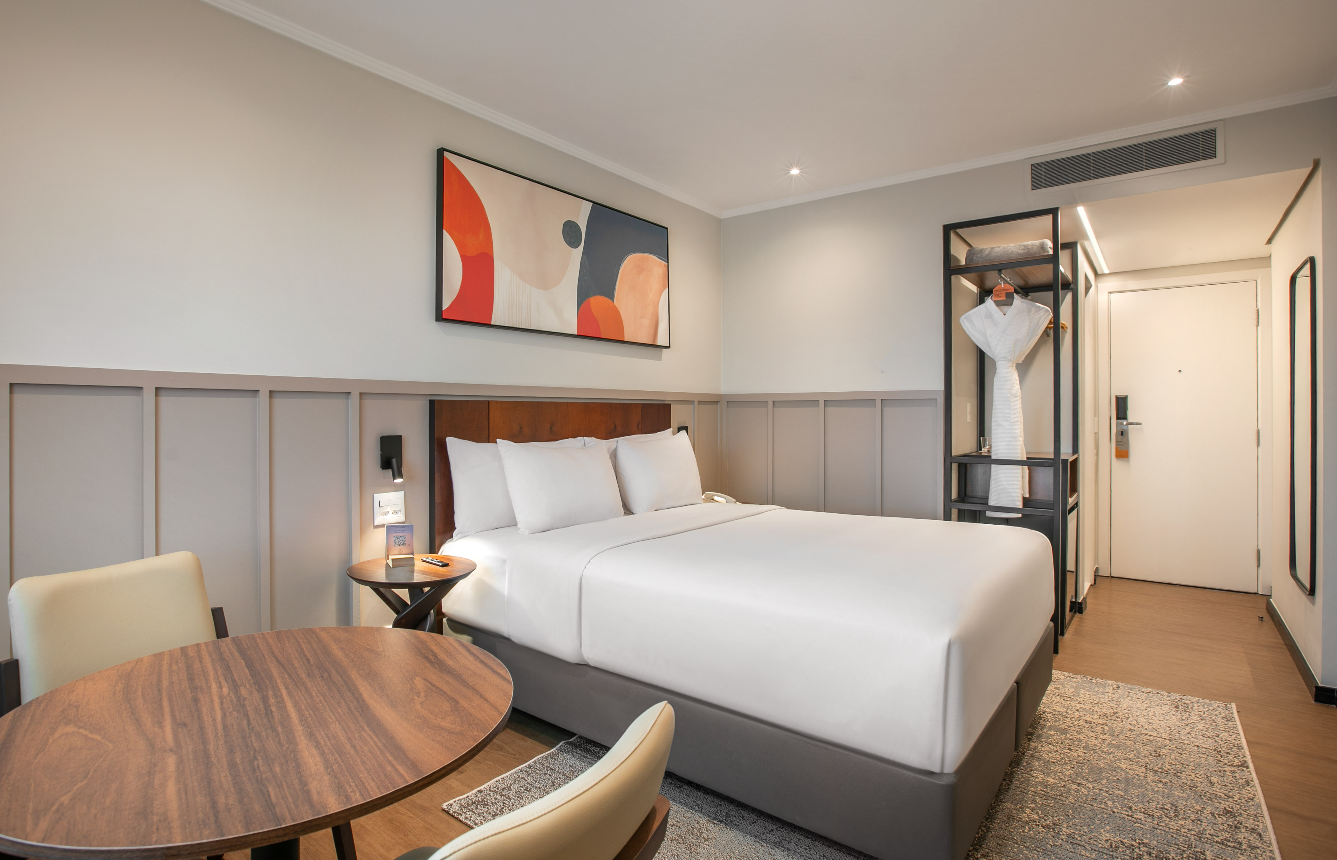 Modern hotel room with bed, abstract art, table, chairs, and open wardrobe.