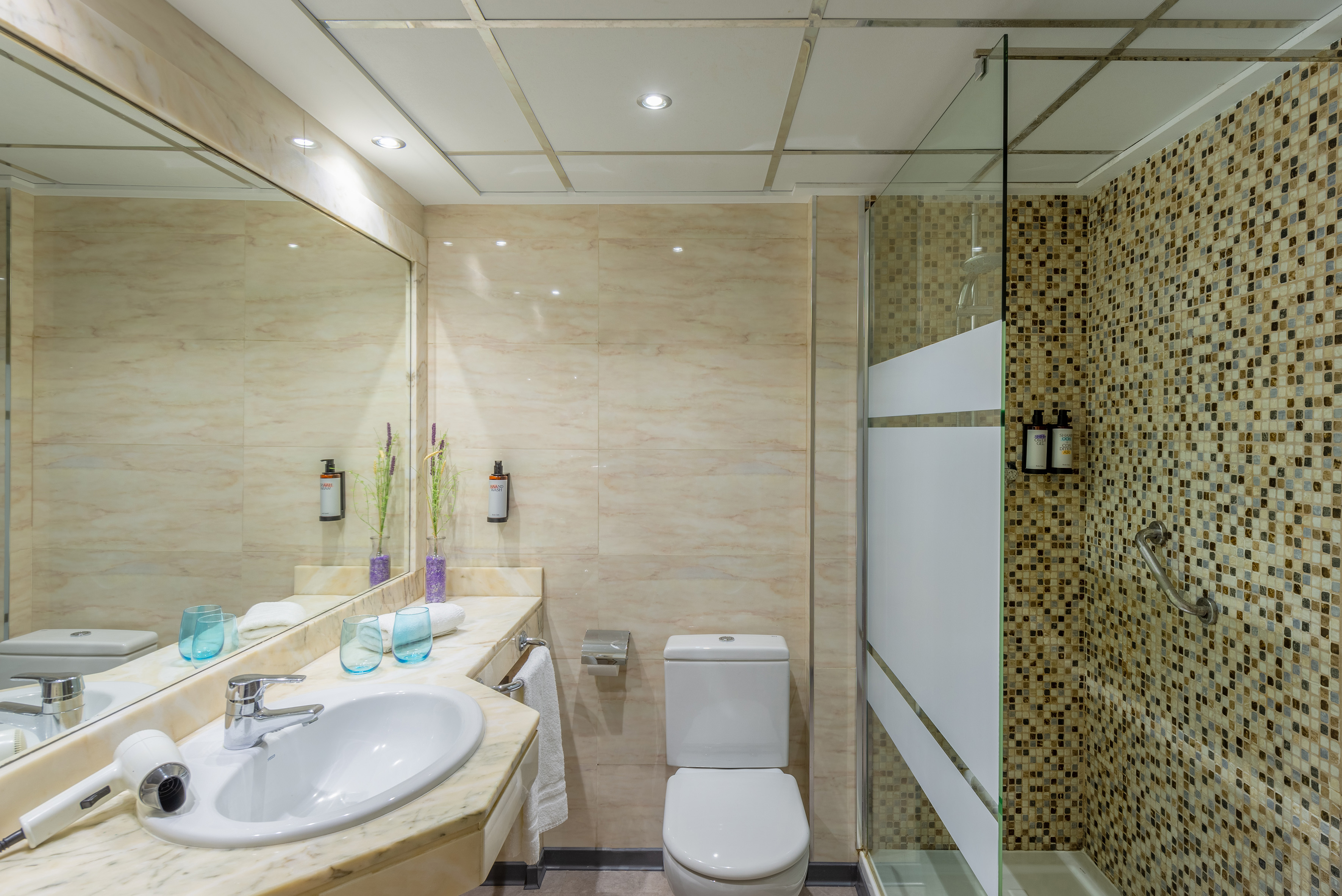 Modern hotel bathroom with marble vanity, sink, toilet, and mosaic tile shower.