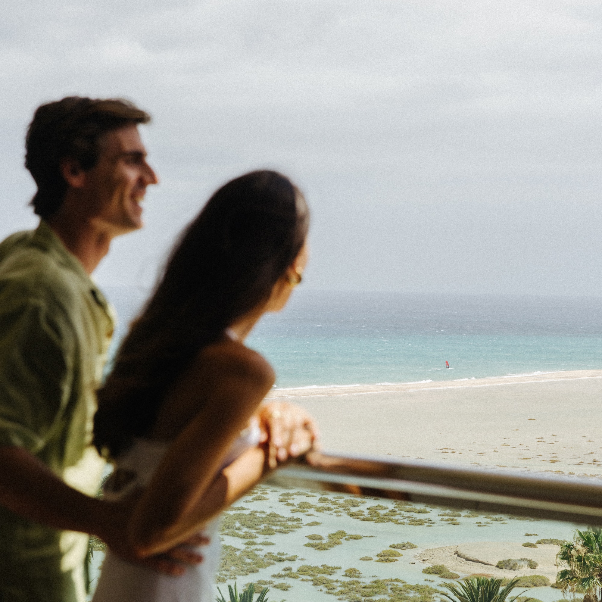 Couple on balcony viewing tropical beach, ocean, and windsurfer.