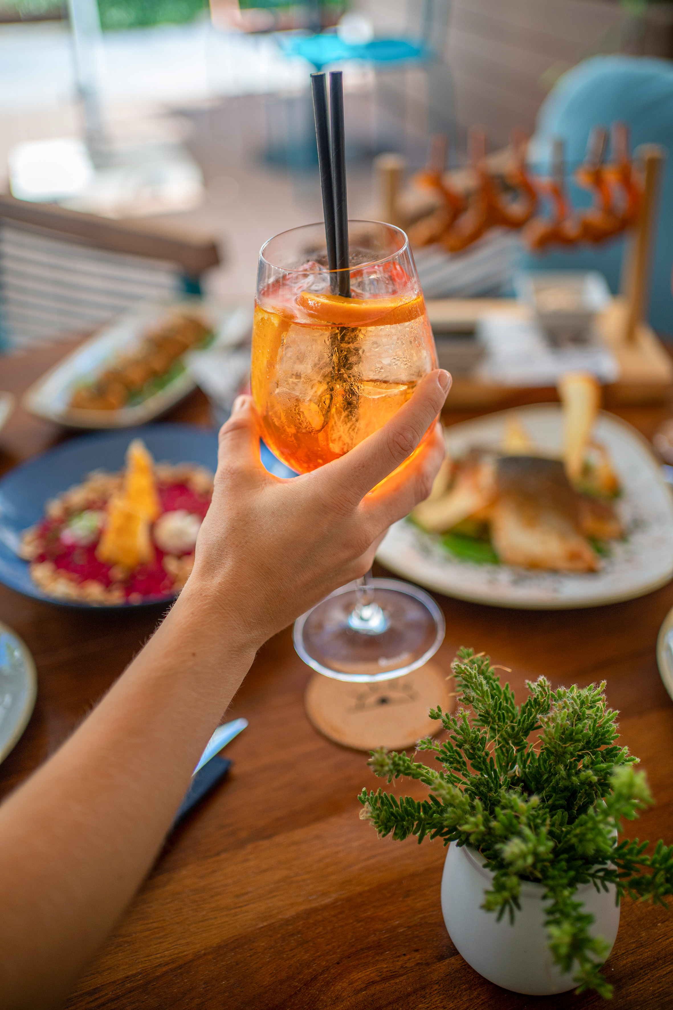 Hand holding an Aperol Spritz cocktail with orange slice and straws at a restaurant table.