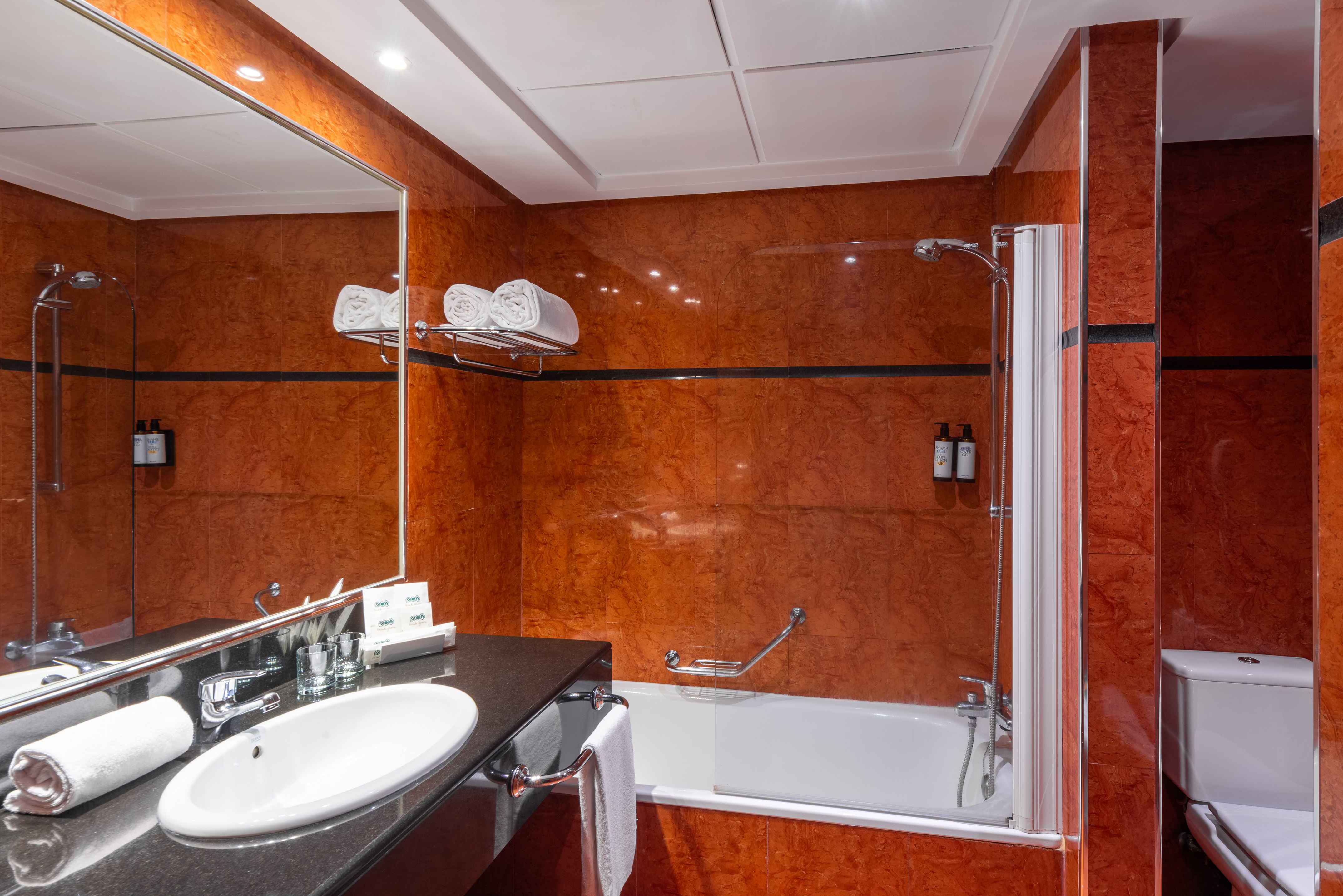 Modern hotel bathroom with red tiled walls, white sink, bathtub, shower, and toilet.