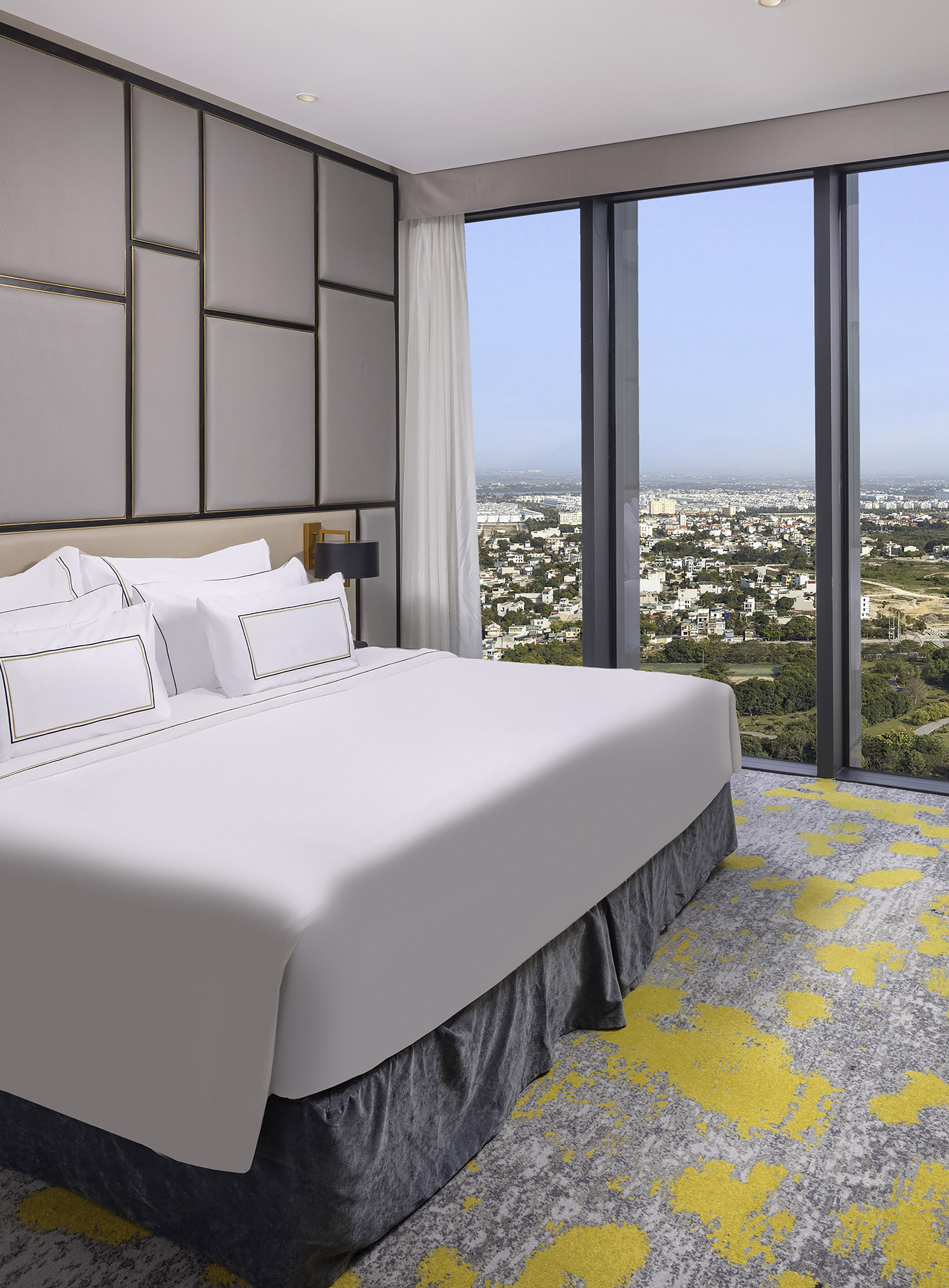 Modern hotel bedroom with city view, king bed, geometric headboard, and abstract carpet.
