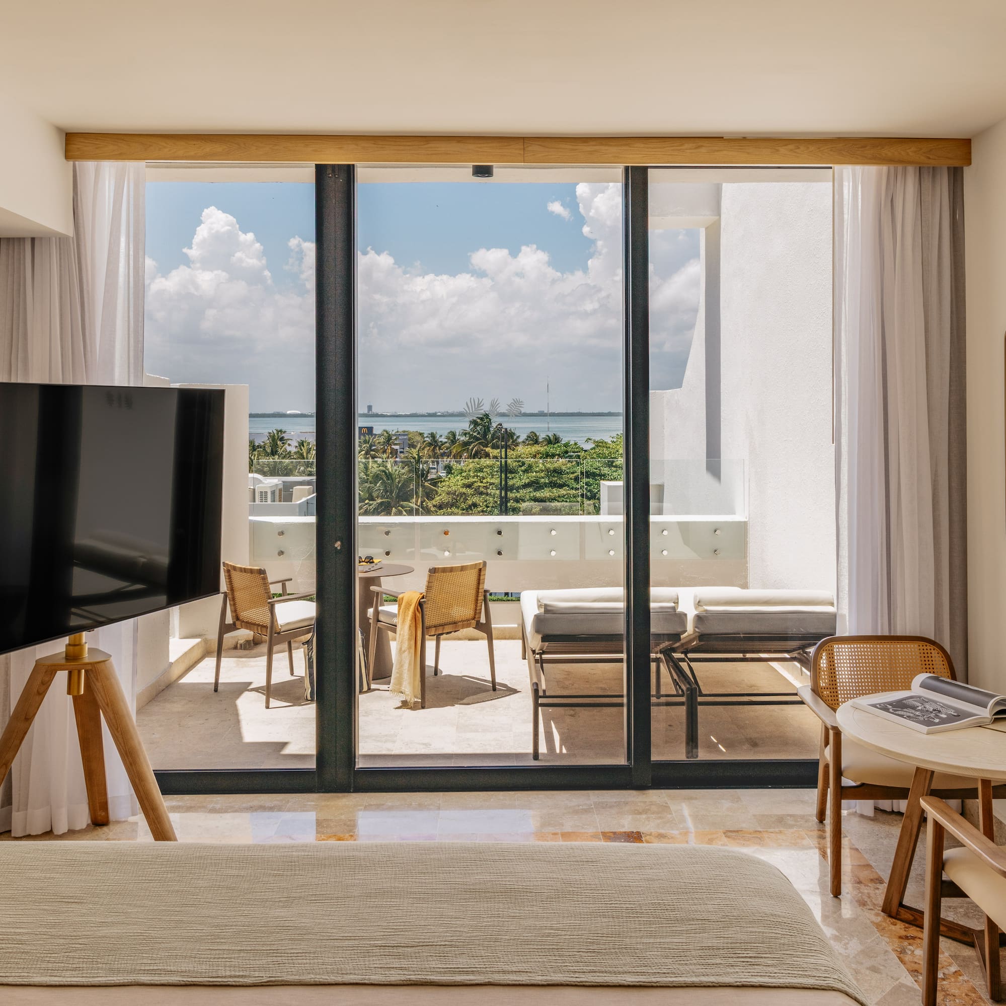Modern hotel room with ocean view balcony, outdoor seating, and lush tropical trees.