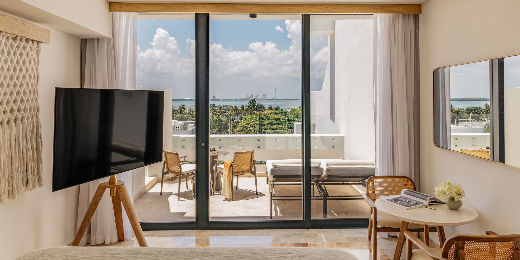 Modern hotel room with ocean view balcony, outdoor seating, and lush tropical trees.