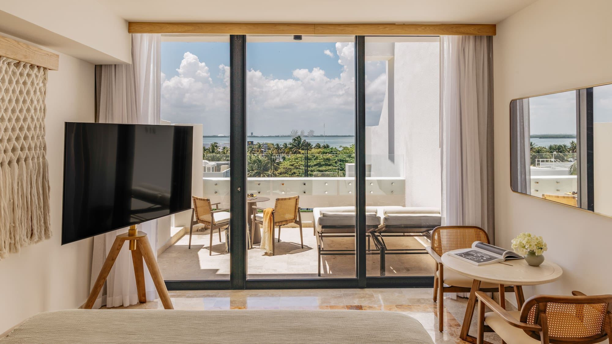 Modern hotel room with ocean view balcony, outdoor seating, and lush tropical trees.