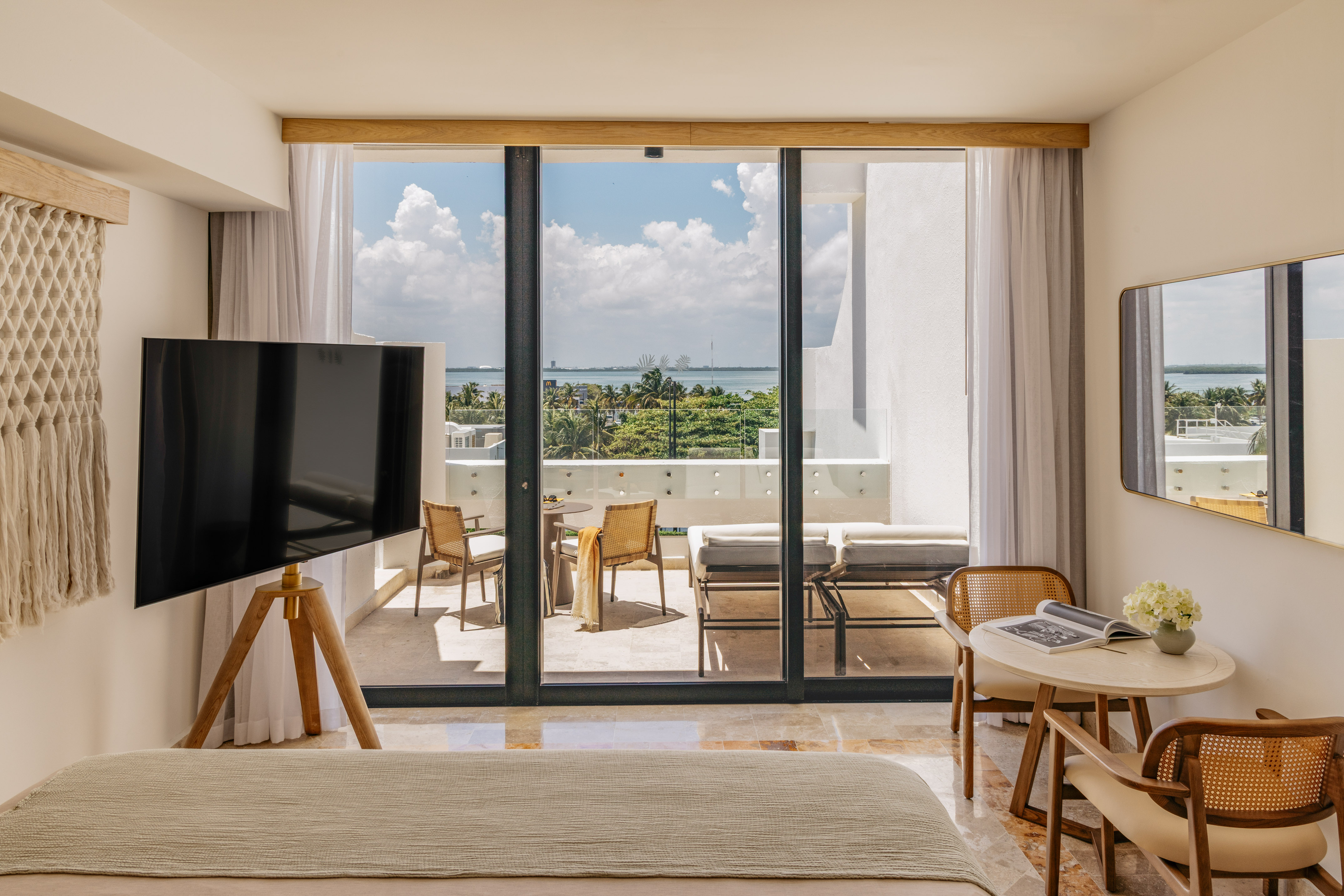 Modern hotel room with ocean view balcony, outdoor seating, and lush tropical trees.