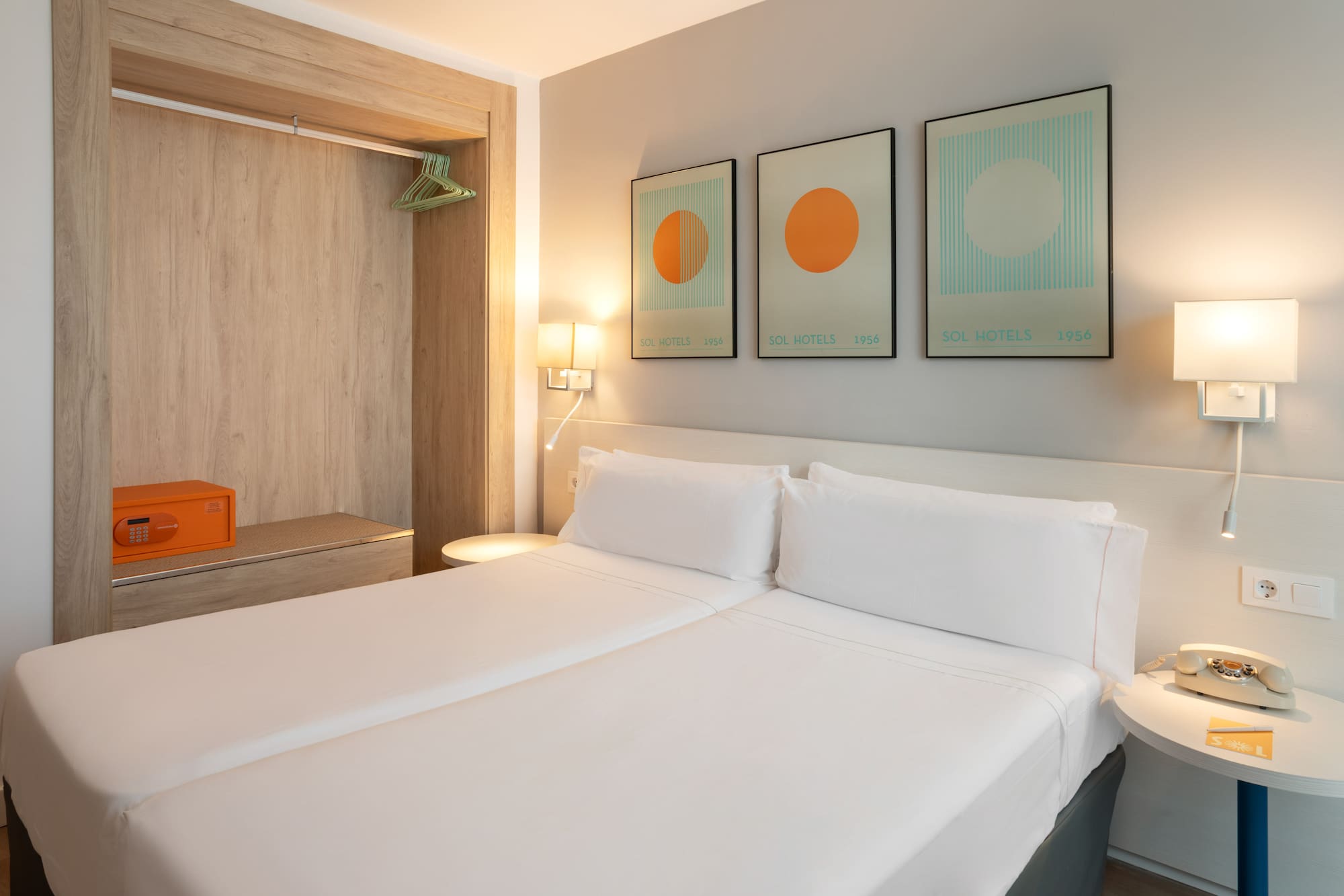 Modern hotel room with twin beds, open wardrobe, orange safe, and geometric wall art.