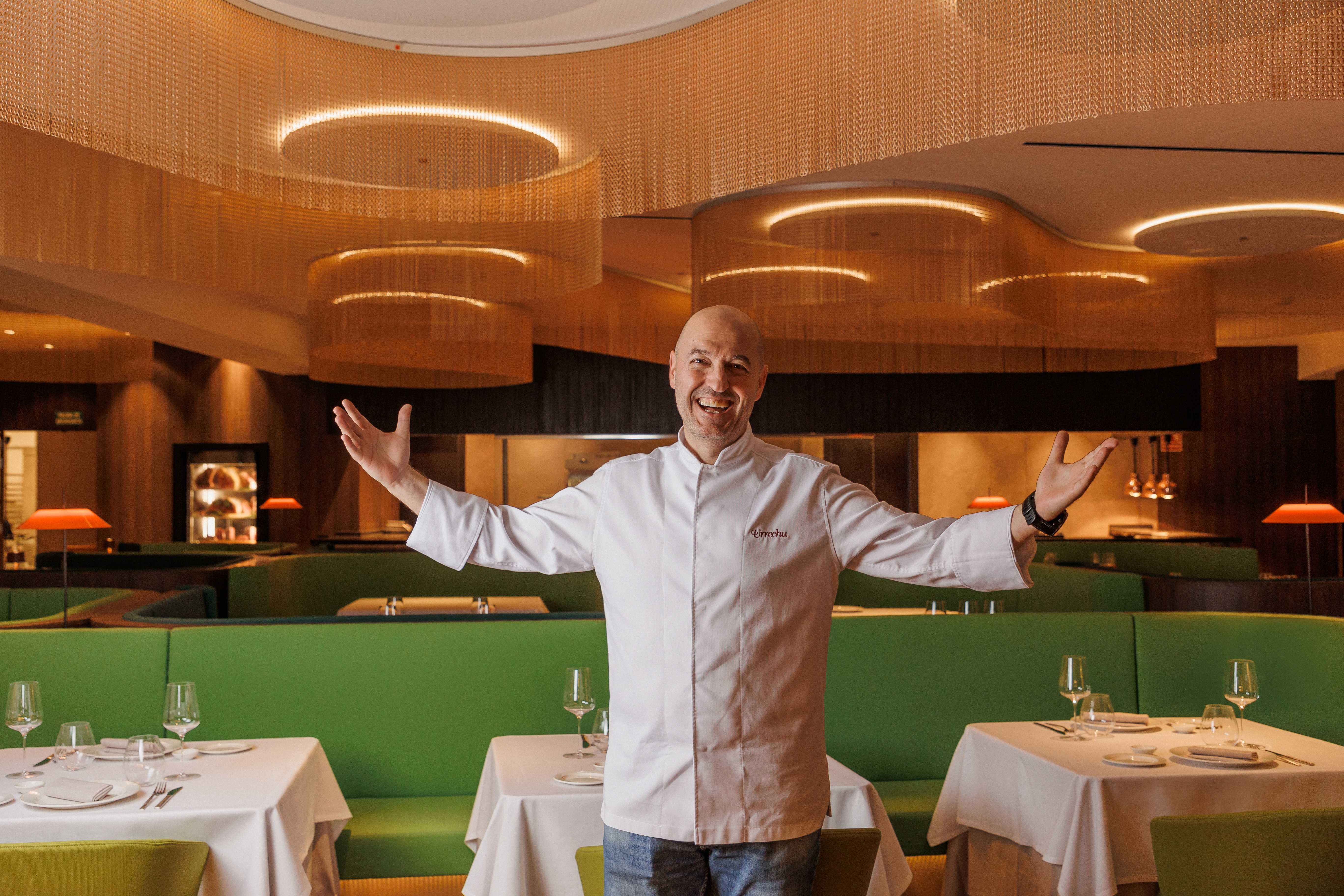 Chef Urrechu, arms open, smiles in his modern restaurant with green banquettes.