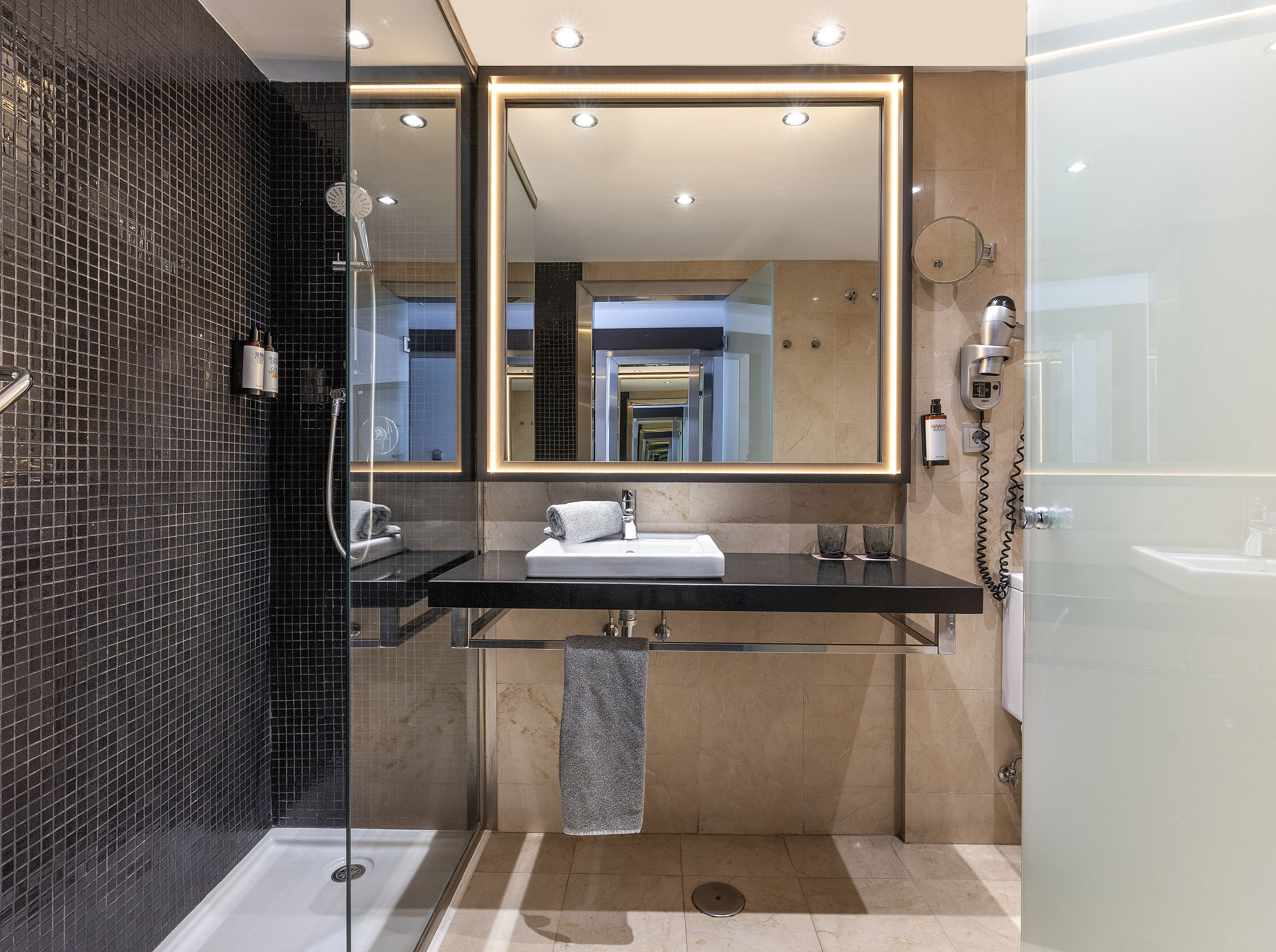 Modern hotel bathroom with black mosaic shower, illuminated mirror, and beige vanity.