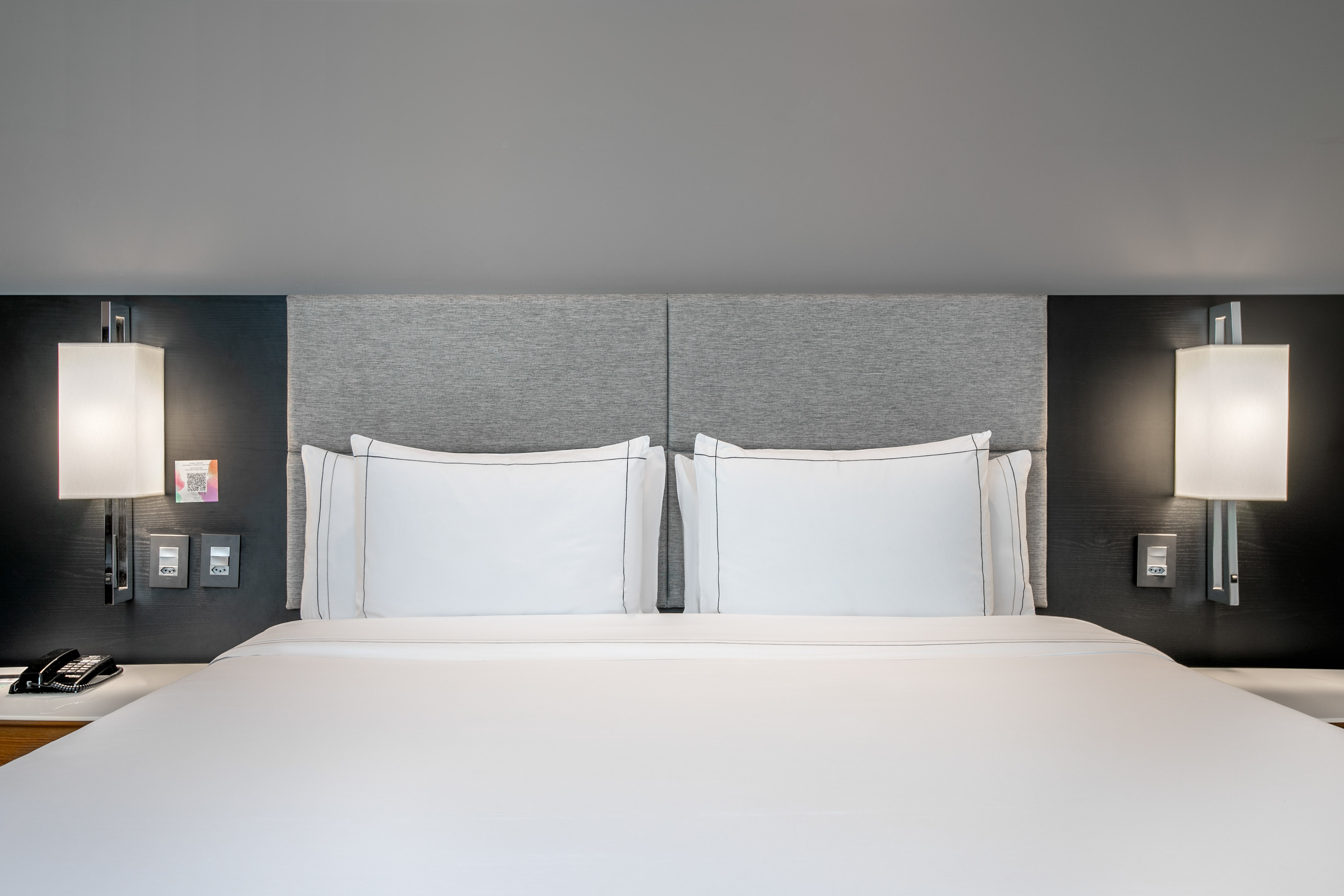 Modern hotel room bed with white linens, grey headboard, and illuminated wall lamps.
