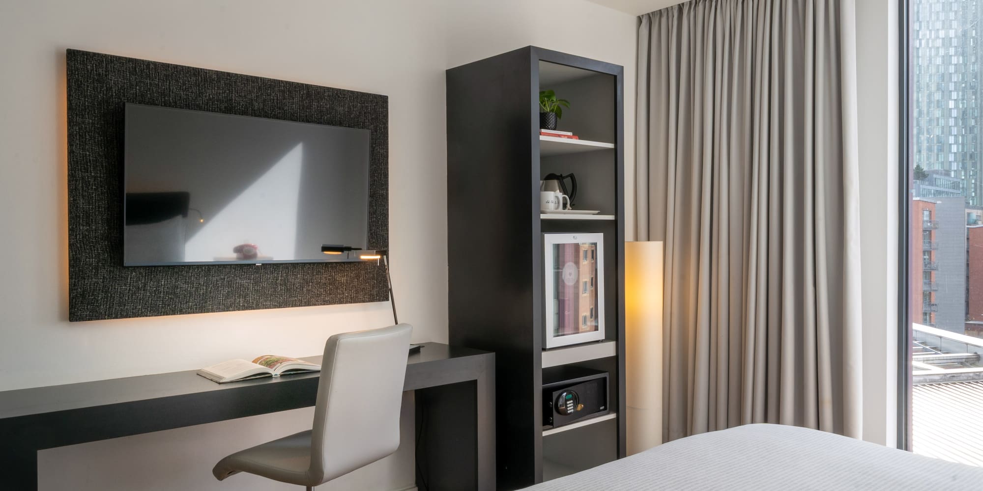 Modern hotel room with desk, TV, mini-fridge, safe, and city view.